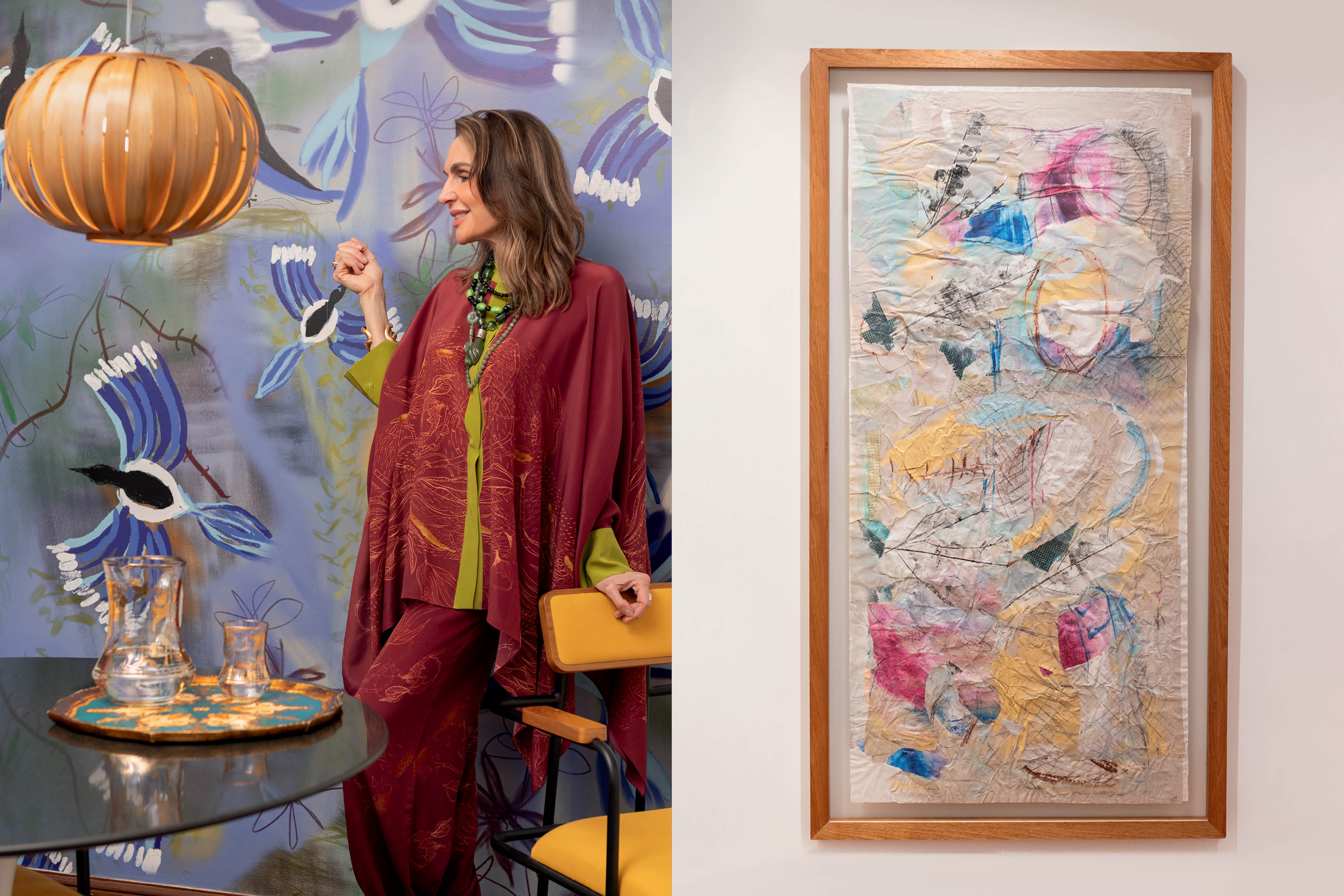 Mulher de cabelos castanhos e blusa vinho com detalhes verdes, sorrindo e olhando para a direita, em ambiente com papel de parede de p&aacute;ssaros azuis e lumin&aacute;ria de madeira. &Agrave; direita, um quadro abstrato com tons pastel e texturas
