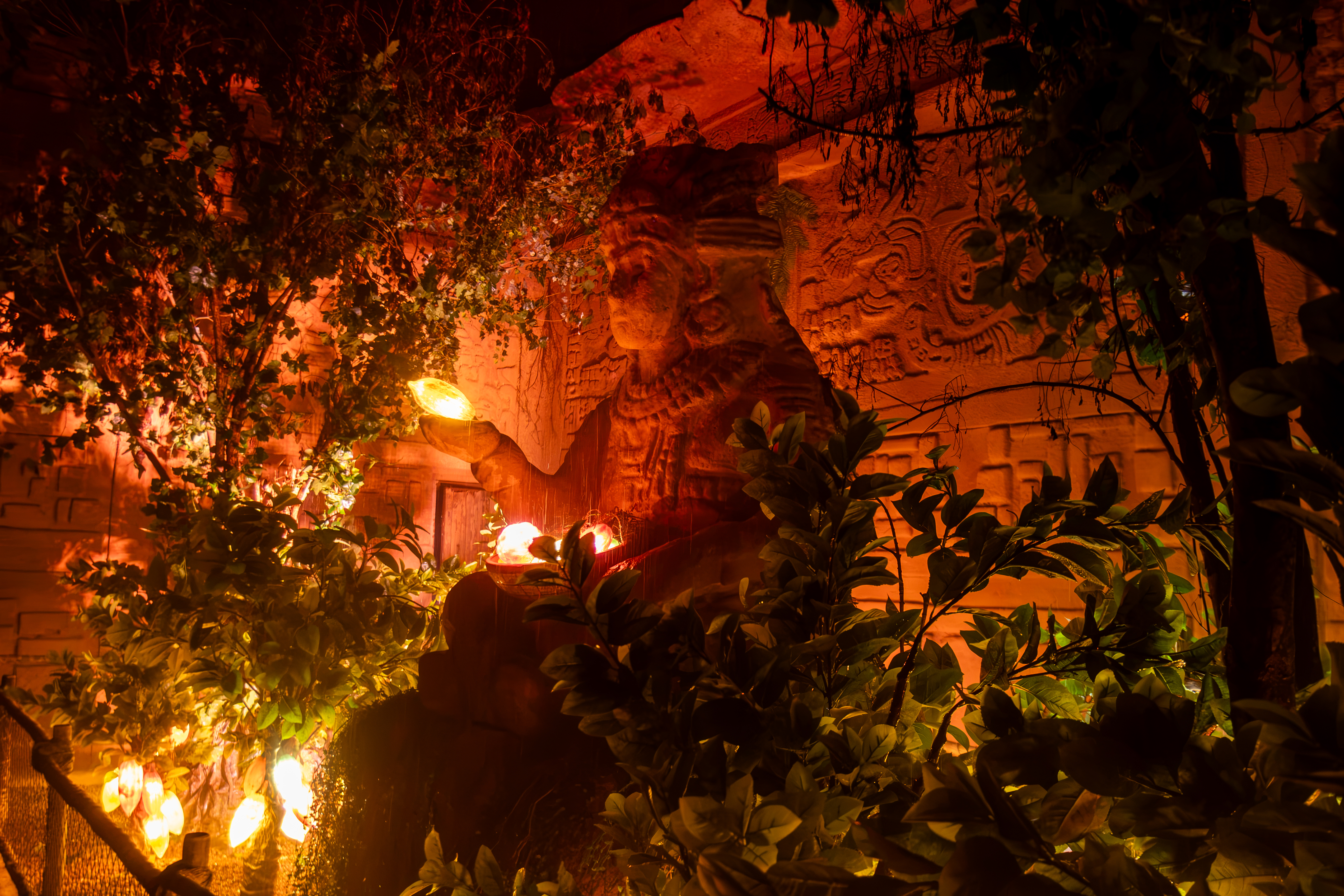 Est&aacute;tua maia de pedra iluminada por luzes laranja e amarelas, com folhagens escuras em primeiro plano e relevos antigos na parede ao fundo