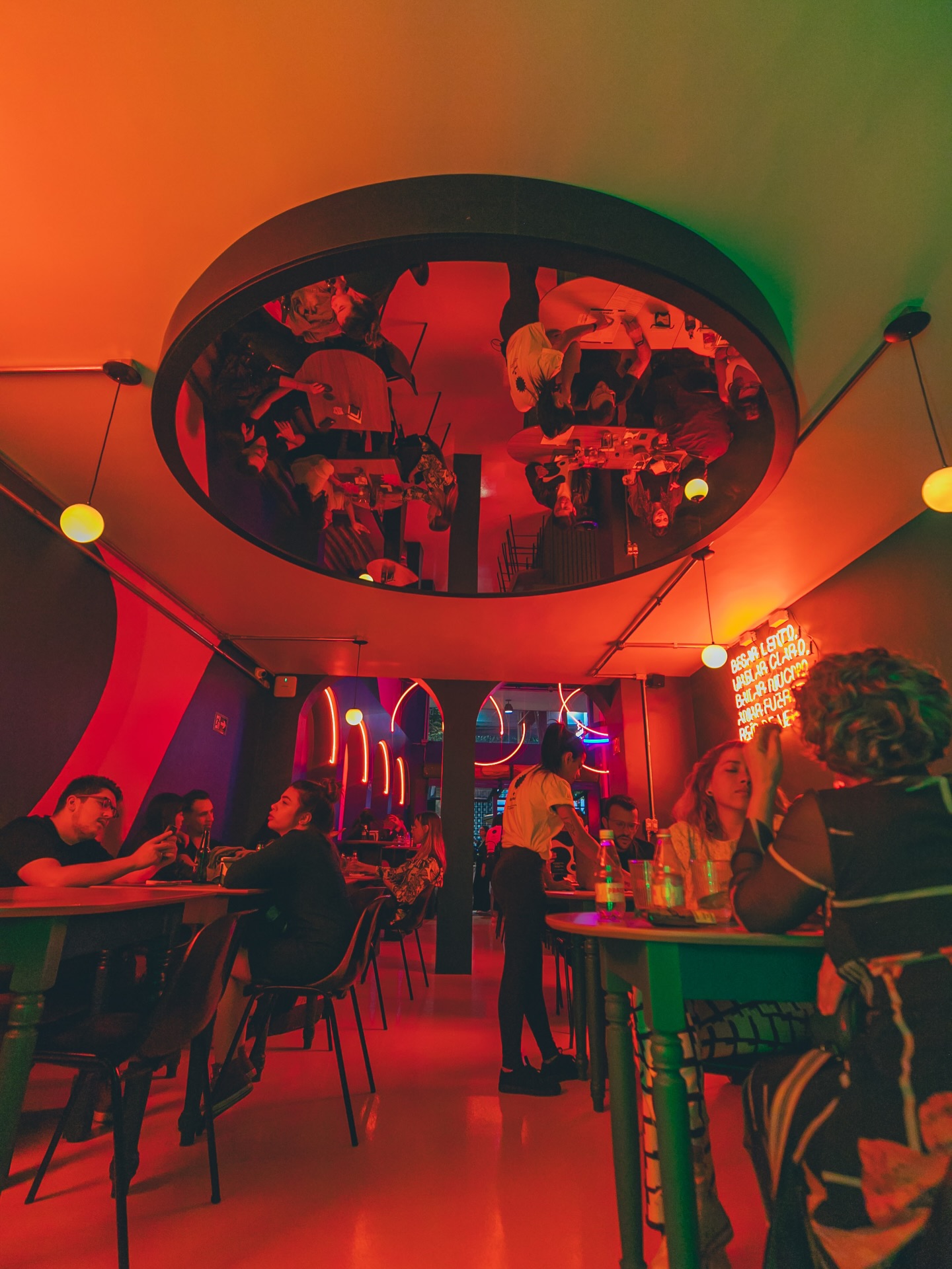 Pessoas em um bar com ilumina&ccedil;&atilde;o neon vermelha e verde, refletidas em um espelho circular no teto. Clientes sentados em mesas de madeira, alguns conversando, outros olhando celulares. Uma gar&ccedil;onete serve uma mesa. O ambiente &eacute; moderno e vibrante
