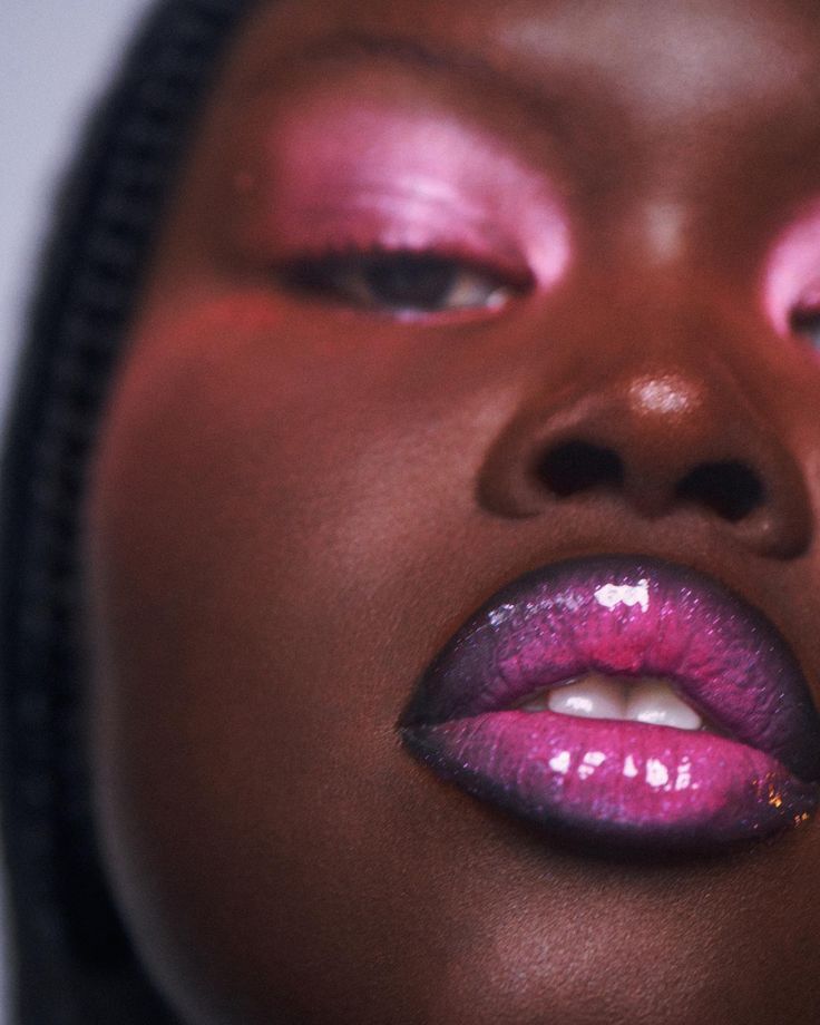 Close-up do rosto de uma pessoa com pele escura, maquiagem vibrante e l&aacute;bios brilhantes. A p&aacute;lpebra superior tem sombra rosa met&aacute;lica, e os l&aacute;bios est&atilde;o pintados em um ombr&eacute; de roxo escuro para rosa choque, com gloss cintilante. Parte do nariz e da bochecha s&atilde;o vis&iacute;veis, com um fundo claro e desfocado