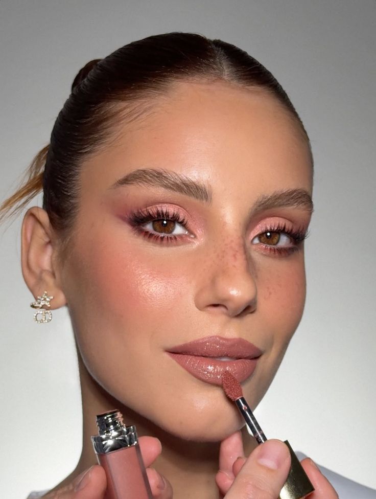 Mulher jovem com sardas, cabelo castanho preso, maquiagem rosa nos olhos e bochechas, aplicando gloss labial nude com um pincel. Ela usa um brinco dourado com as letras CD