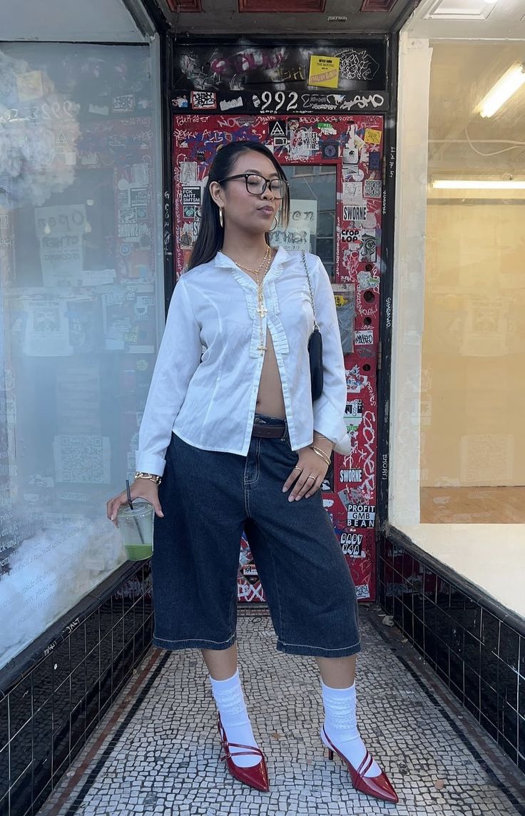 Mulher asi&aacute;tica com &oacute;culos, camisa branca aberta, bermuda jeans e sapatos vermelhos, segurando um copo verde, em frente a uma vitrine coberta de adesivos e grafites