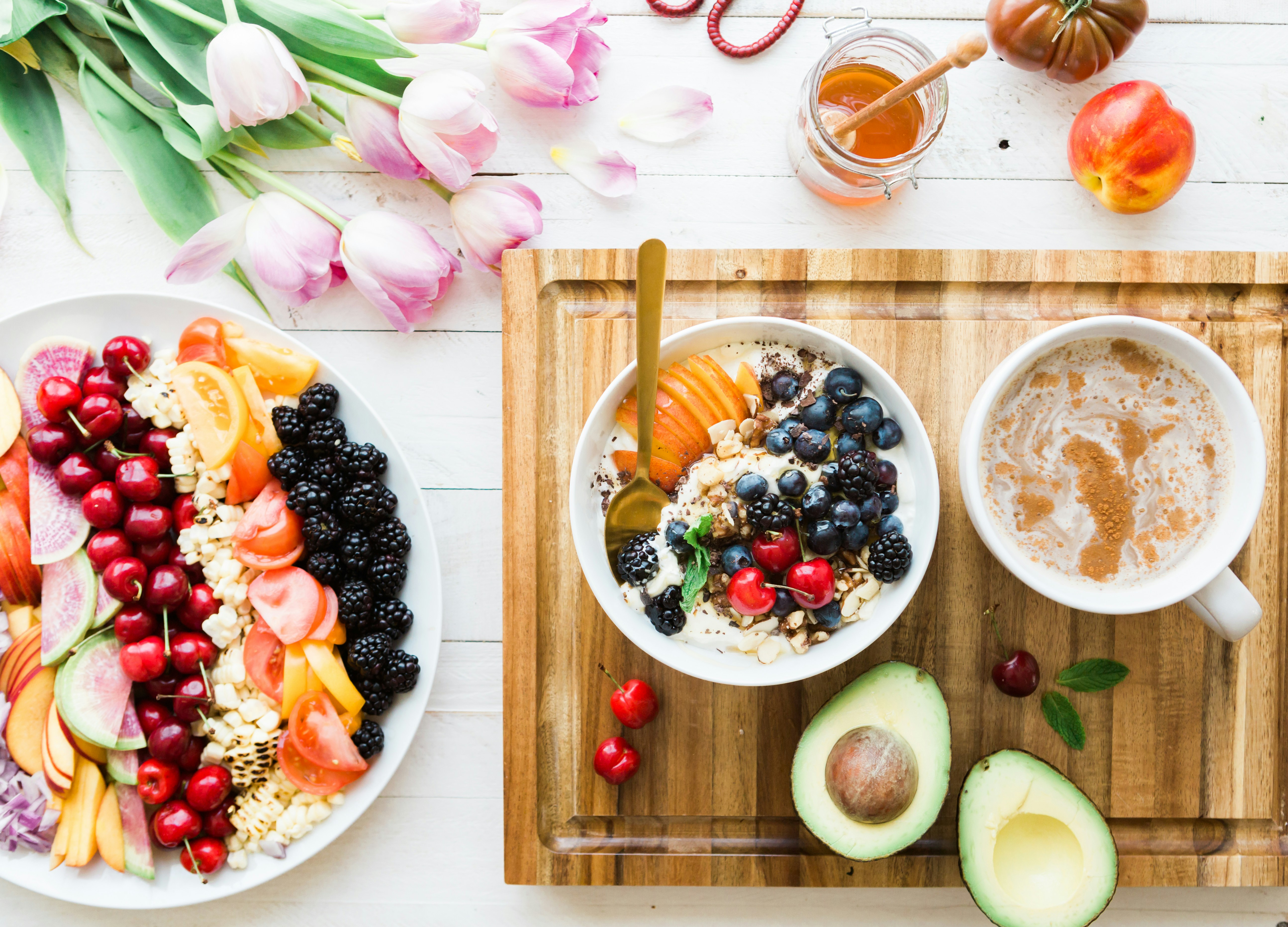 Mesa de madeira clara com um prato oval de frutas variadas, um bowl de iogurte com frutas e uma xícara de café com canela. Ao fundo, tulipas cor-de-rosa e um pote de mel.