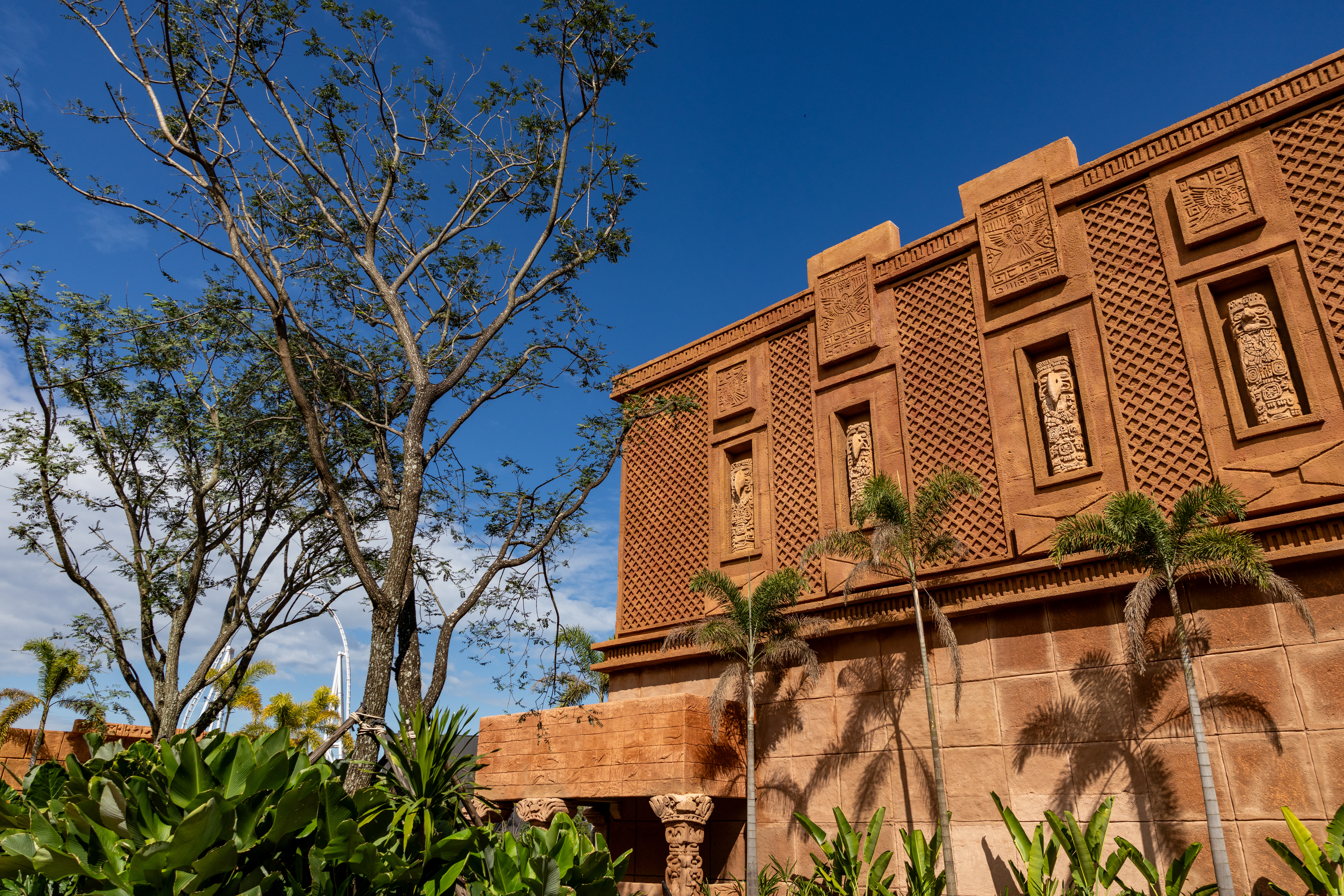 Edif&iacute;cio de cor terracota com detalhes em relevo de estilo maia, palmeiras e folhagens verdes em primeiro plano, sob c&eacute;u azul com nuvens esparsas