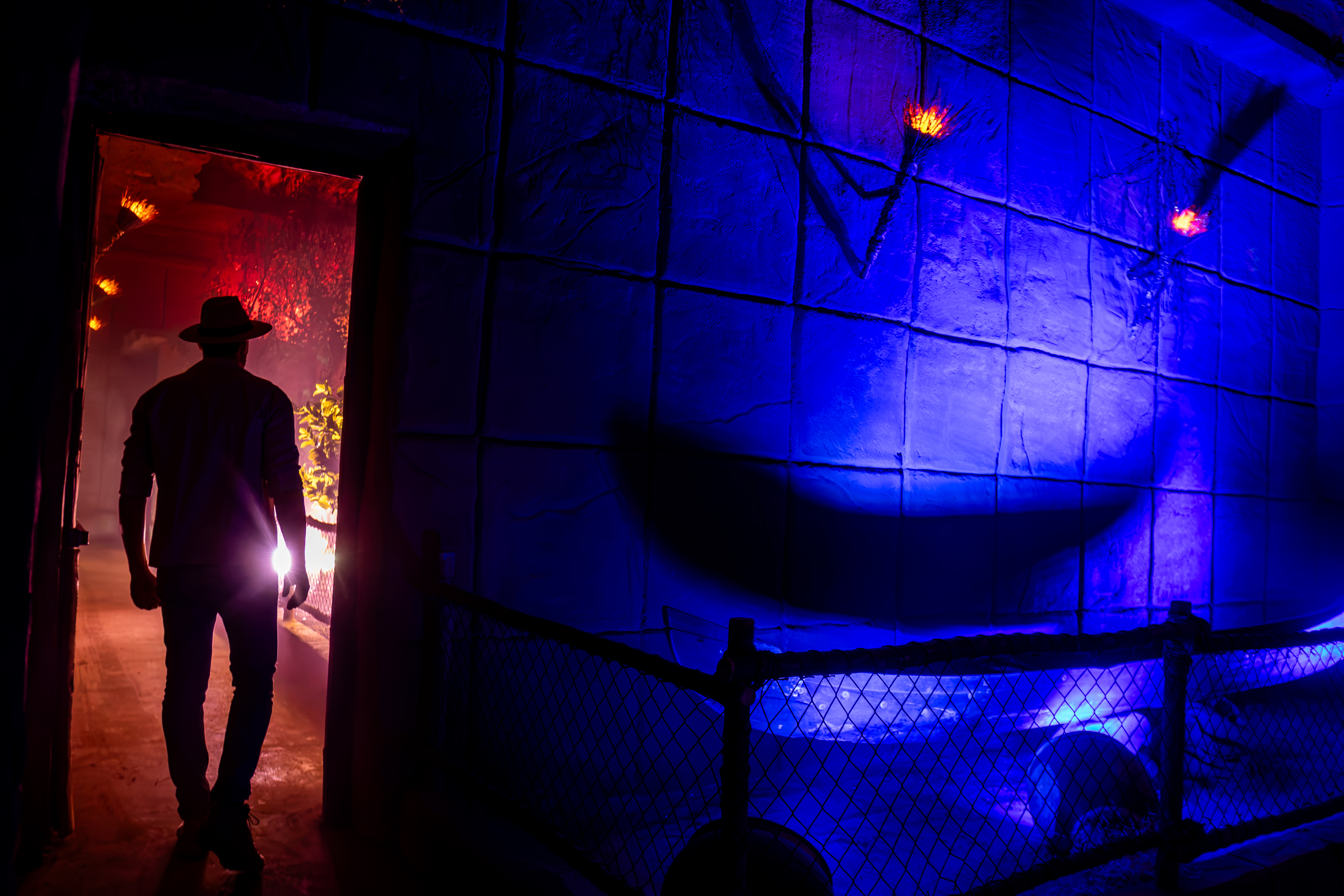 Homem de chap&eacute;u e camisa escura, de costas, caminha por um corredor iluminado em tons de vermelho e laranja, com folhagens &agrave; direita. &Agrave; frente, uma parede azul com luzes e uma cerca de tela preta.