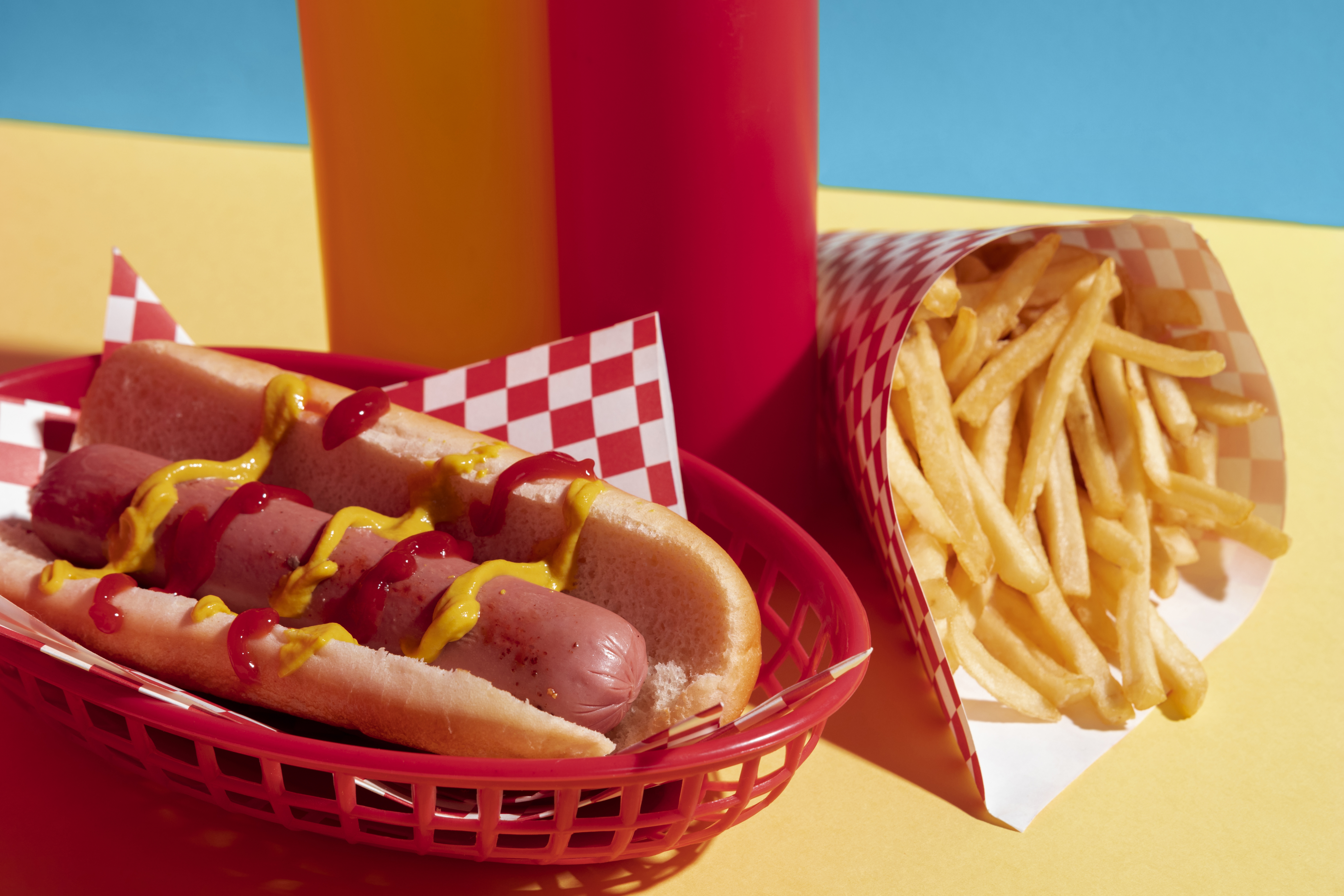Cachorro-quente com mostarda e ketchup em cesta vermelha, batatas fritas em cone de papel xadrez, e garrafas de molho ao fundo, sobre fundo amarelo e azul