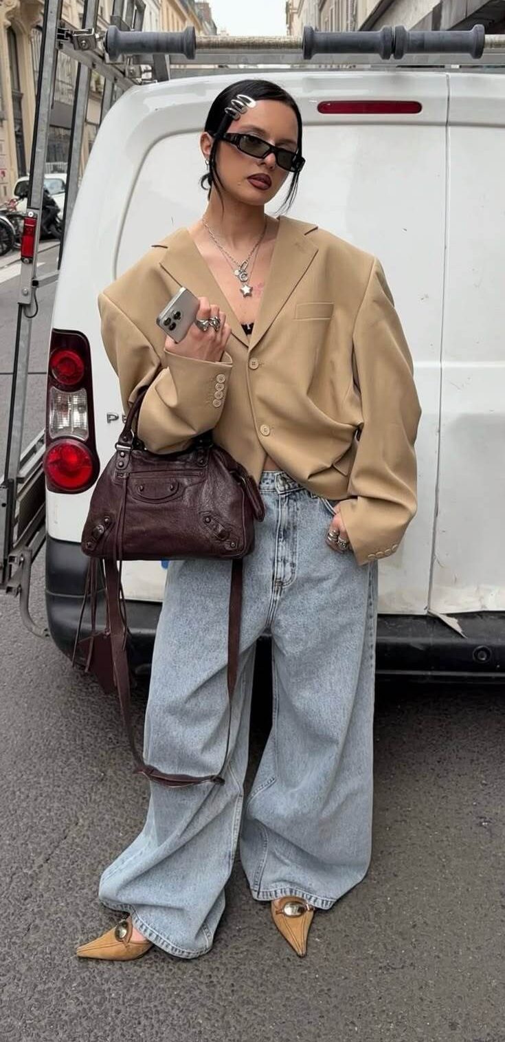 Mulher com &oacute;culos escuros, blazer bege oversized, cal&ccedil;a jeans larga, bolsa marrom e sapatos de bico fino, posando em frente a uma van branca na rua