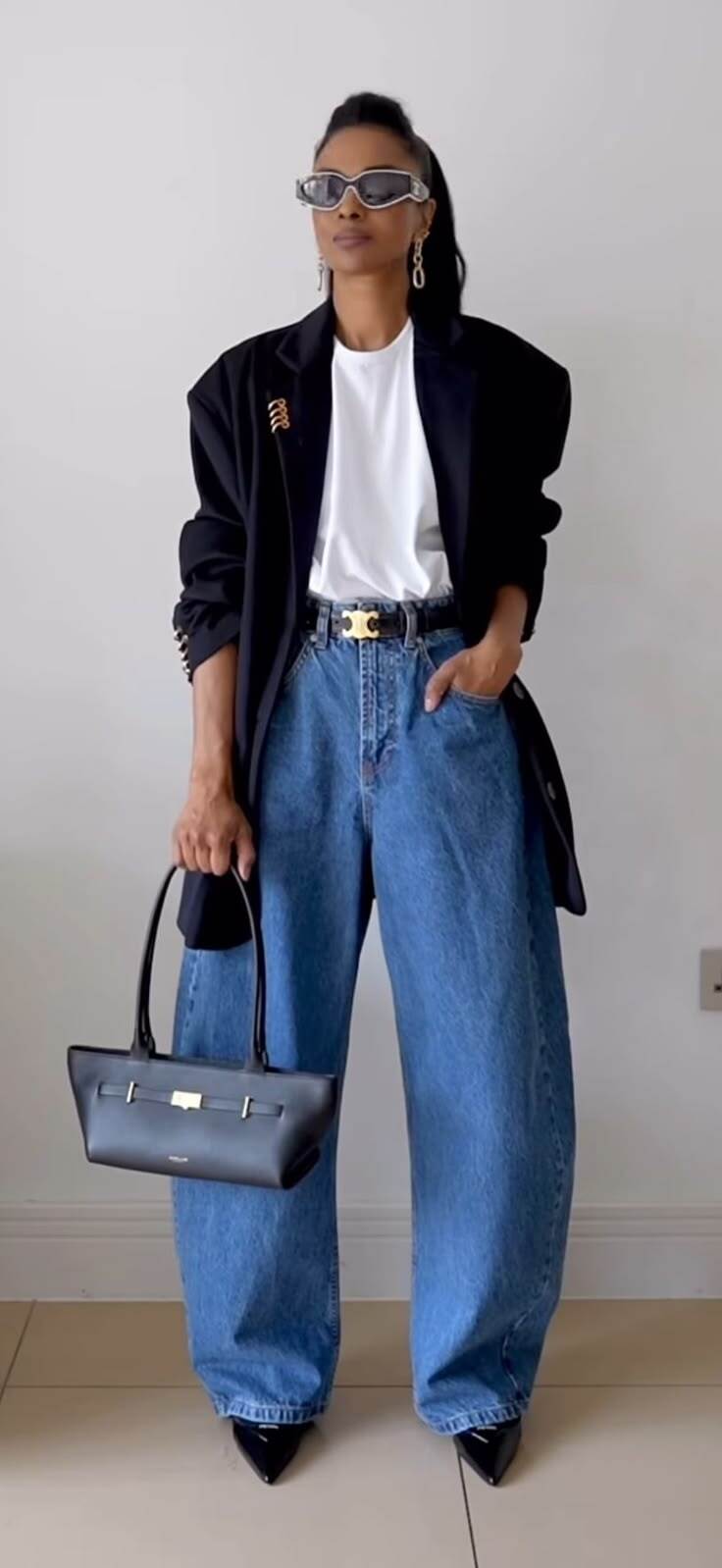 Mulher de pele morena, cabelo escuro preso em rabo de cavalo, usando &oacute;culos de sol futuristas, blazer preto, camiseta branca, cal&ccedil;a jeans larga, cinto preto com fivela dourada e bolsa preta de al&ccedil;a curta, posando com a m&atilde;o no bolso