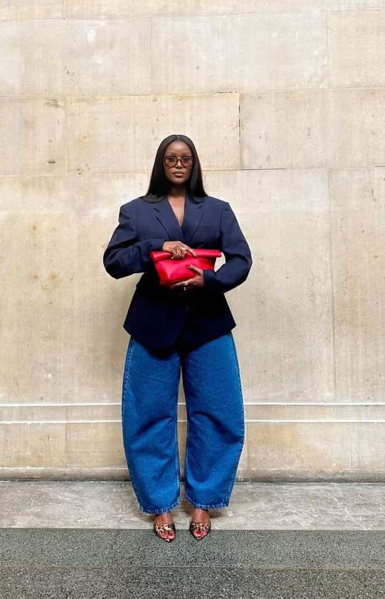 Mulher negra de &oacute;culos escuros, blazer azul-marinho e cal&ccedil;a jeans larga, segurando uma bolsa clutch vermelha, posa em frente a uma parede de pedra clara