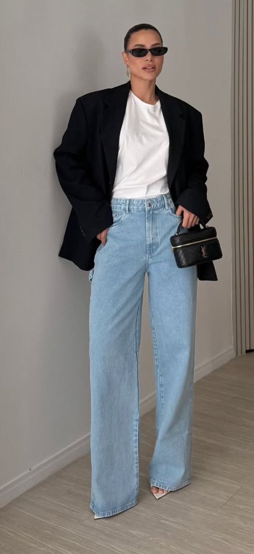 Mulher com cabelo preso, &oacute;culos escuros, blazer preto, camiseta branca, cal&ccedil;a jeans larga e bolsa preta na m&atilde;o, posando em p&eacute; contra uma parede cinza