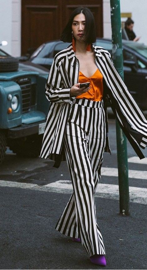 Mulher asi&aacute;tica de cabelo preto curto, usando um conjunto listrado preto e branco com blusa laranja e sapatos roxos, caminhando na rua e segurando um celular
