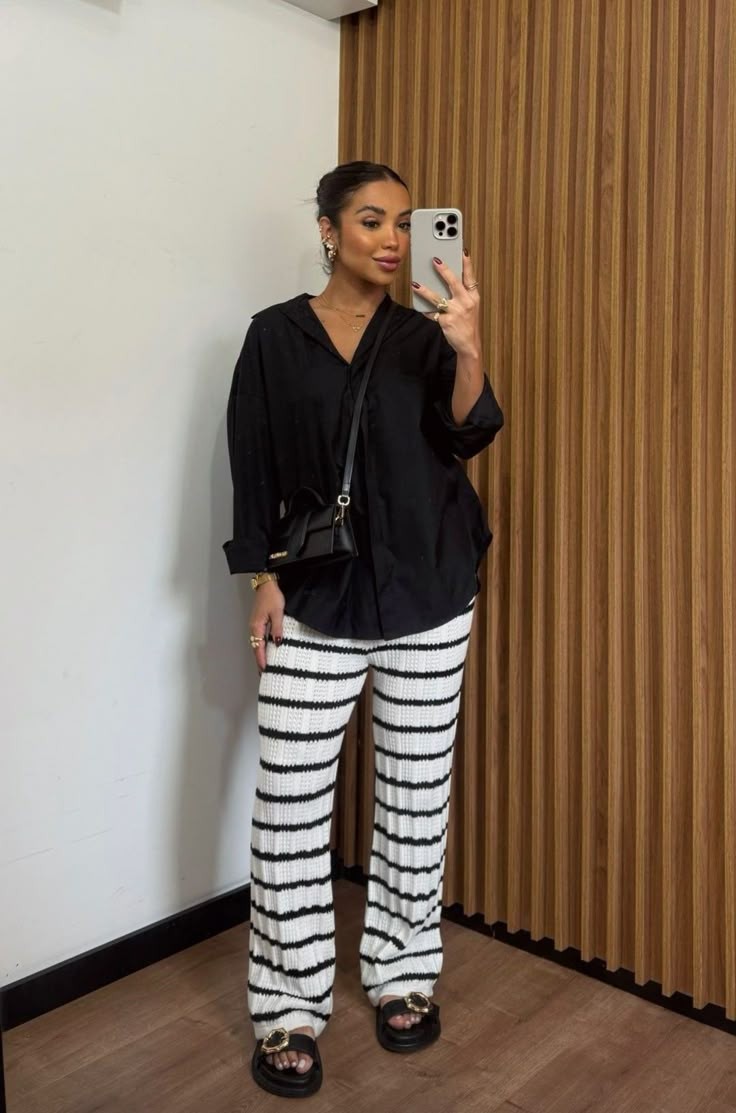 Mulher sorridente de pele morena e cabelo preso, vestindo camisa preta folgada, cal&ccedil;a de tric&ocirc; branca com listras pretas horizontais e sand&aacute;lias pretas. Ela segura um celular para tirar uma selfie, exibindo an&eacute;is e brincos dourados. Uma bolsa preta pequena atravessa seu corpo. O fundo &eacute; uma parede branca e outra de madeira ripada