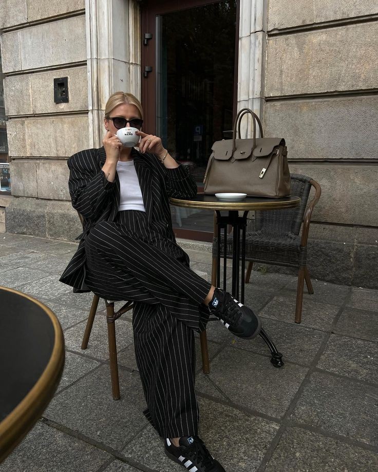 Mulher loira de &oacute;culos escuros, blazer e cal&ccedil;a listrados pretos, sentada em uma cadeira de caf&eacute;, bebendo de uma x&iacute;cara branca. Uma bolsa marrom est&aacute; na mesa ao lado, em um ambiente externo com piso de pedra e fachada de pr&eacute;dio.