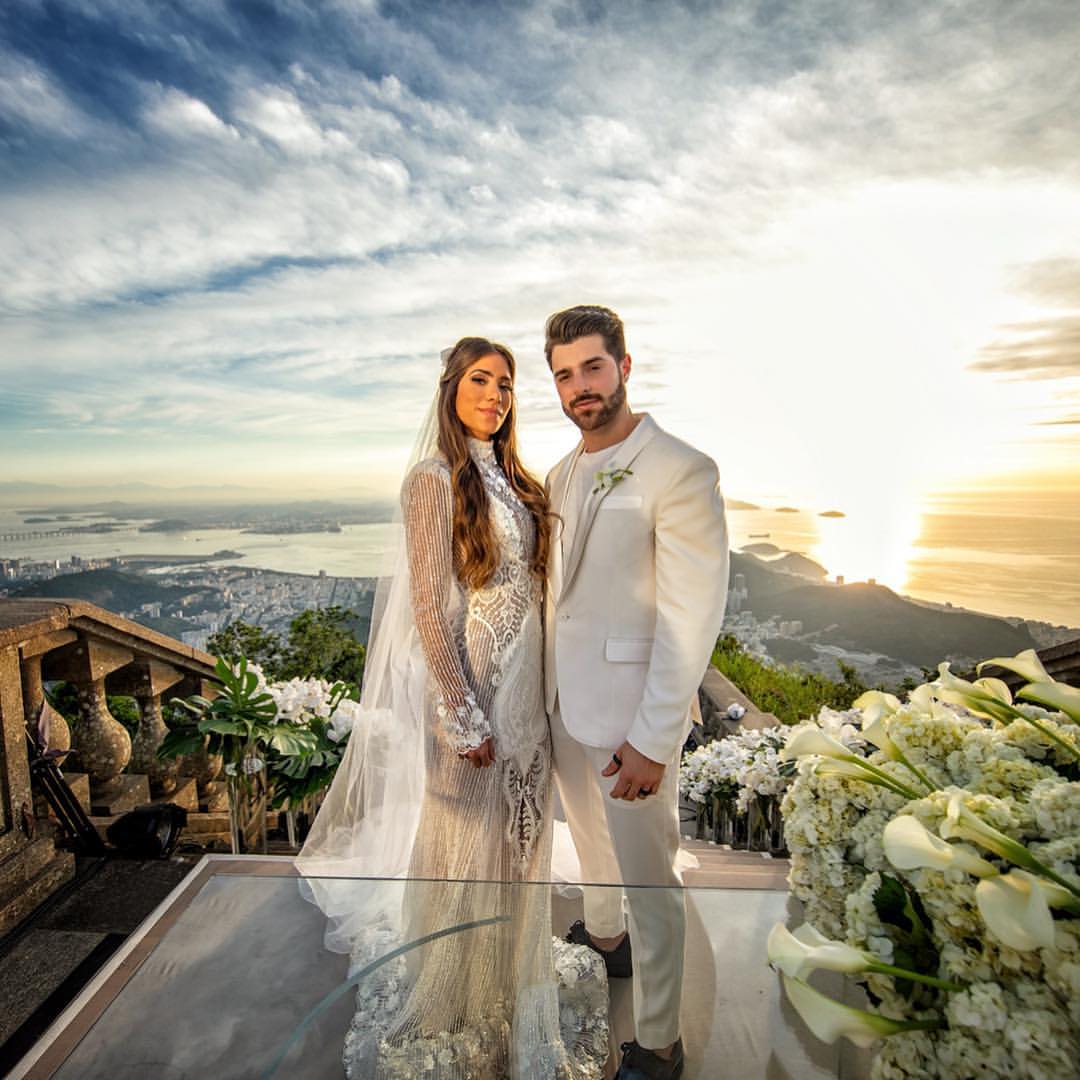 Casal em trajes de casamento, a mulher com vestido de renda e v&eacute;u, o homem de terno branco, posam em um mirante com vista para o mar e a cidade ao p&ocirc;r do sol, com arranjos de flores brancas ao redor