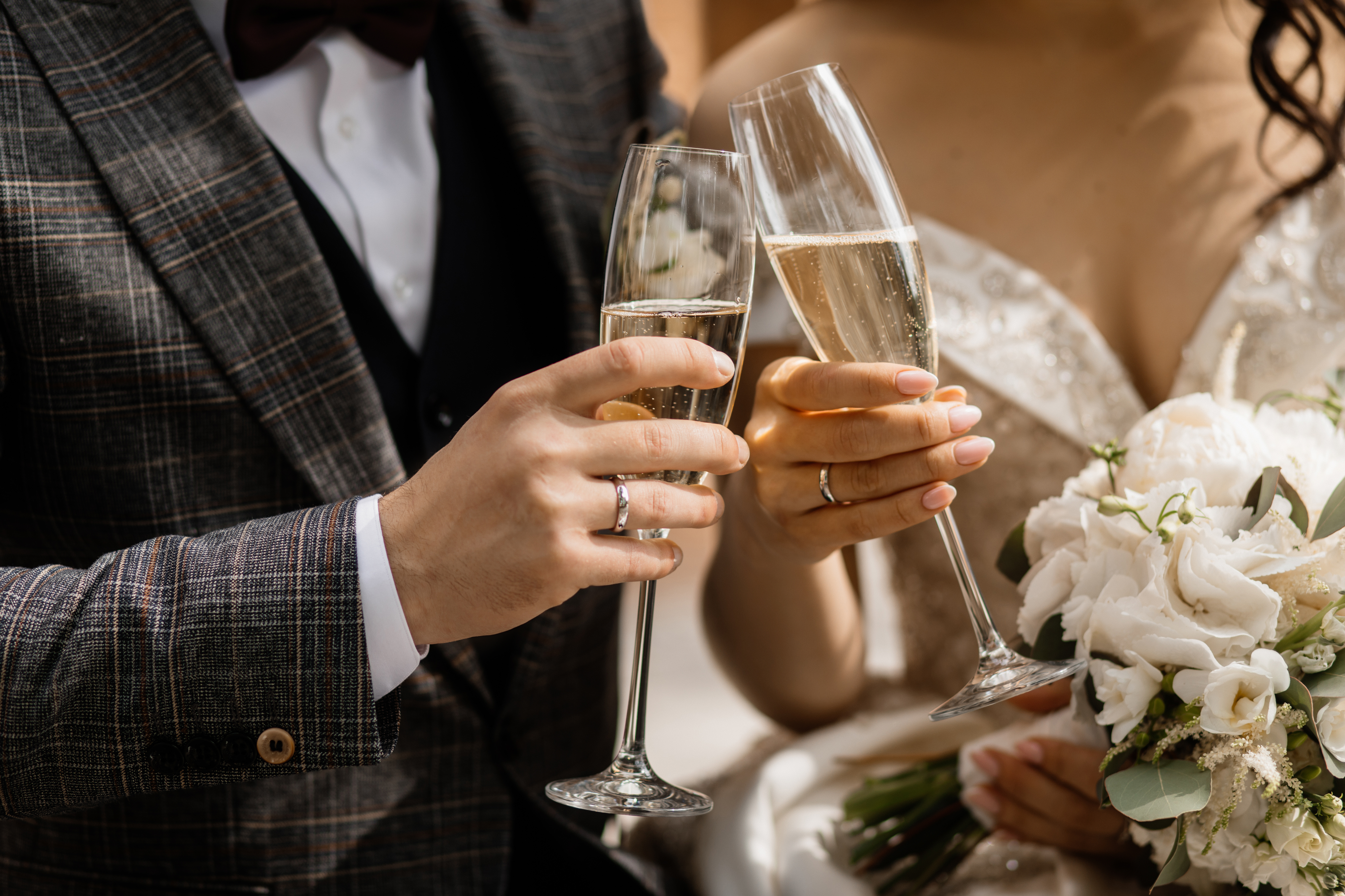 Um casal brinda com ta&ccedil;as de champanhe, mostrando suas m&atilde;os com alian&ccedil;as. O homem veste um terno xadrez marrom e a mulher um vestido de noiva branco com um buqu&ecirc; de flores brancas