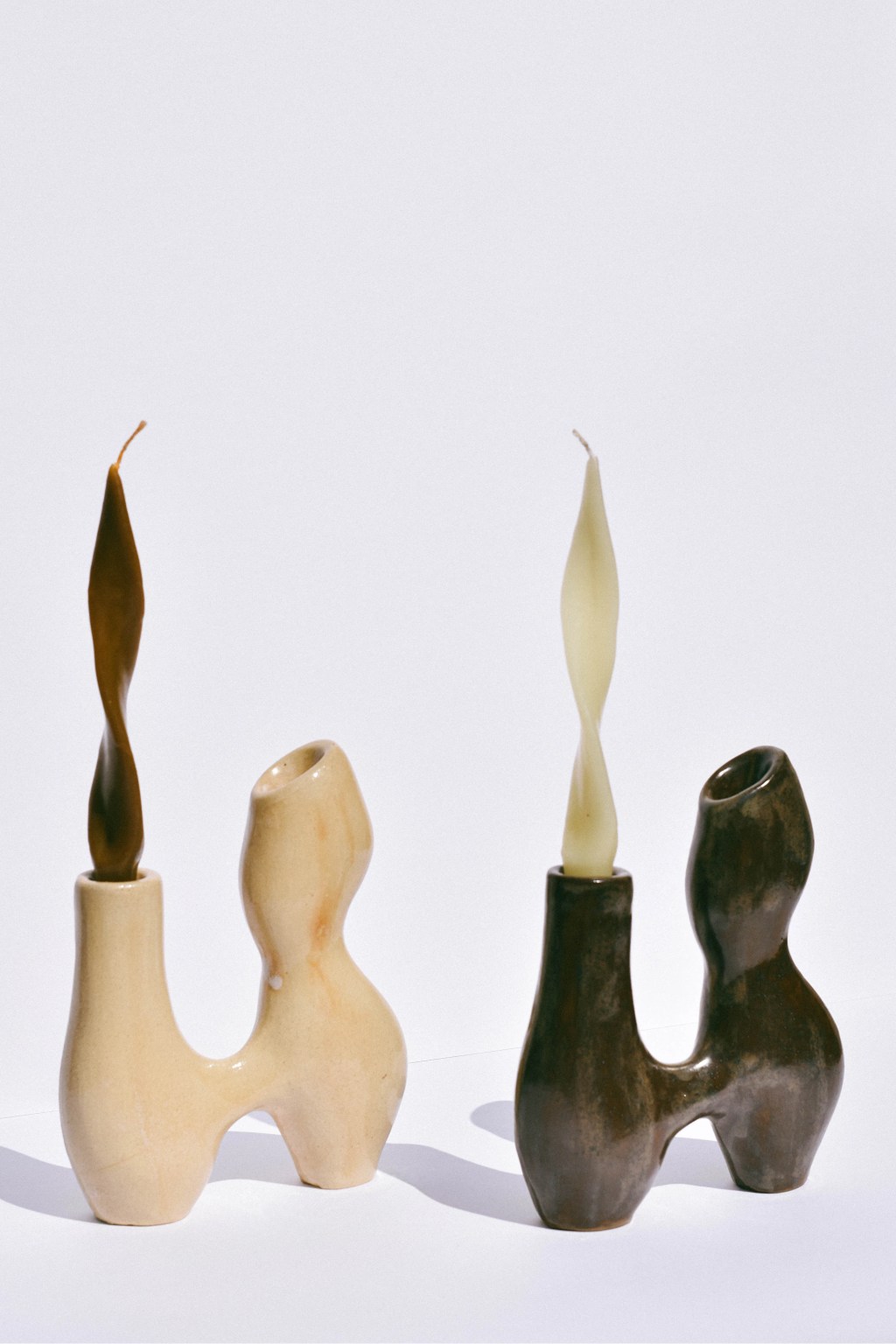 Dois casti&ccedil;ais de cer&acirc;mica com velas torcidas em um fundo branco. O casti&ccedil;al esquerdo &eacute; bege claro com uma vela marrom escura. O casti&ccedil;al direito &eacute; marrom escuro com uma vela bege clara. Ambos t&ecirc;m formato org&acirc;nico, lembrando silhuetas humanas.