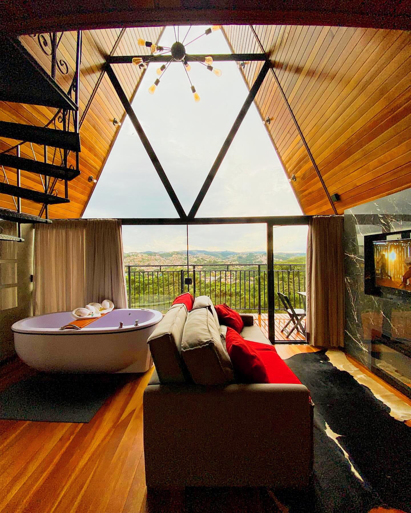 Interior de um loft moderno com banheira lil&aacute;s, sof&aacute; cinza com almofadas vermelhas e piso de madeira. Uma grande janela triangular revela uma vista panor&acirc;mica de uma cidade verdejante e montanhas ao fundo. Uma escada em espiral preta e um lustre industrial pendem do teto de madeira. Uma TV est&aacute; embutida em uma parede de m&aacute;rmore preto.