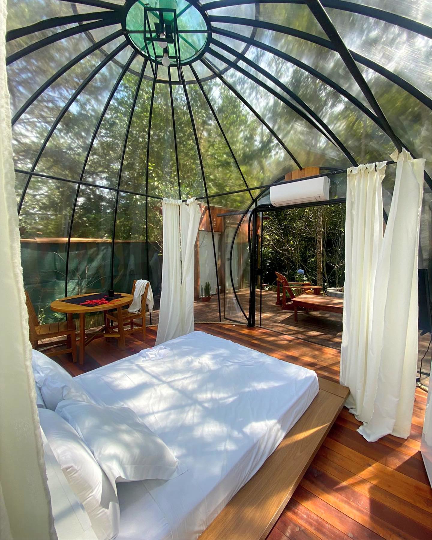 Quarto em formato de domo com teto e paredes de vidro, cercado por &aacute;rvores. Uma cama de casal com len&ccedil;&oacute;is brancos ocupa o centro, e &agrave; esquerda, uma mesa redonda de madeira com duas cadeiras, decorada com p&eacute;talas vermelhas. Cortinas brancas pendem do teto, e um ar-condicionado est&aacute; na parede. O ch&atilde;o &eacute; de madeira, e uma porta de vidro leva a um deck externo com espregui&ccedil;adeira