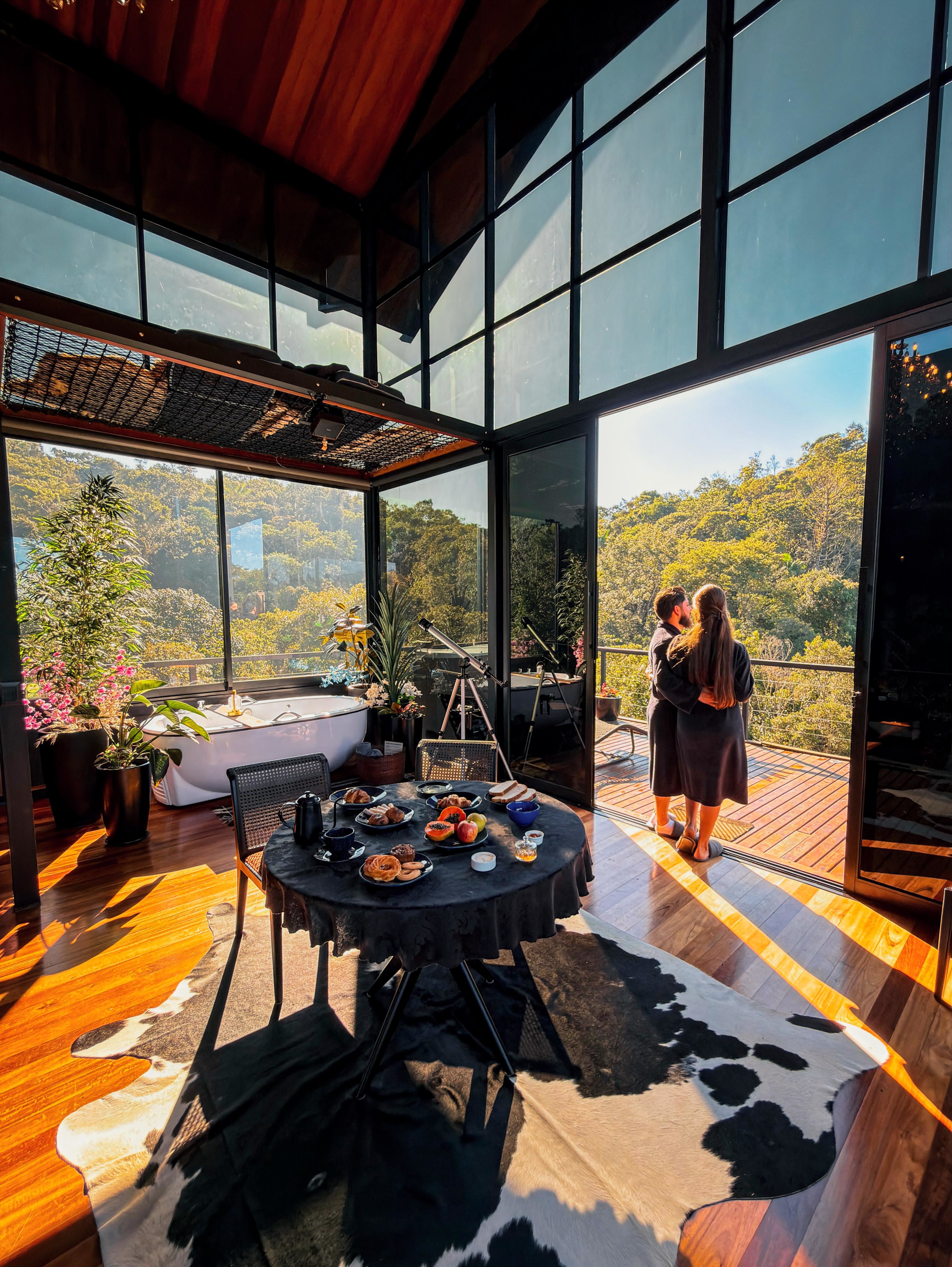 Um casal abra&ccedil;ado, de costas, em uma varanda de madeira com vista para uma floresta exuberante. Dentro do quarto, uma mesa redonda com caf&eacute; da manh&atilde;, uma banheira branca e um telesc&oacute;pio, tudo sob um teto de madeira e grandes janelas que refletem o sol