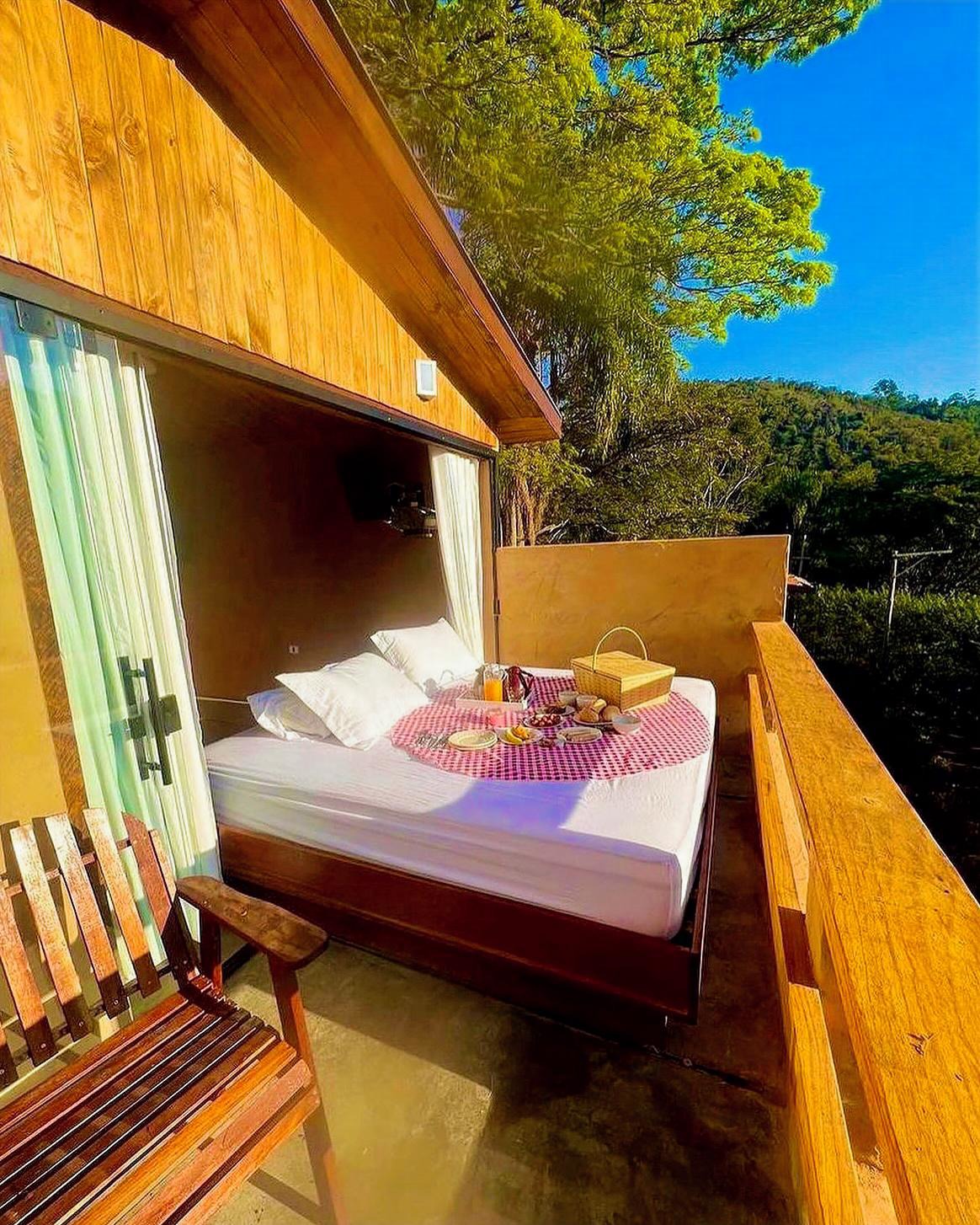 Cama de casal com len&ccedil;&oacute;is brancos e uma toalha xadrez vermelha e branca, com caf&eacute; da manh&atilde; servido em uma bandeja, em uma varanda de madeira com vista para uma paisagem montanhosa e c&eacute;u azul