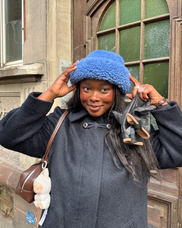 Mulher negra sorridente, com tran&ccedil;as longas, usando um chap&eacute;u azul felpudo e casaco preto. Ela segura luvas de couro pretas e uma bolsa marrom com um chaveiro de ovelha, em frente a uma porta de madeira antiga com vidro verde