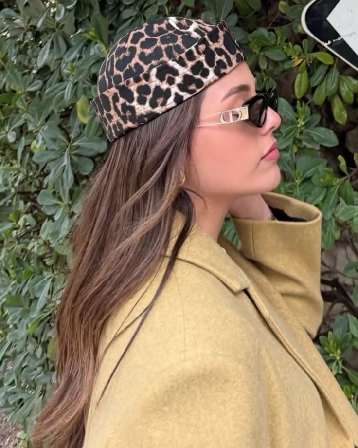 Mulher jovem de perfil, com cabelos castanhos longos, usando um gorro de estampa animal print, &oacute;culos de sol pretos com hastes douradas e um casaco mostarda, em frente a uma parede de folhagens verdes