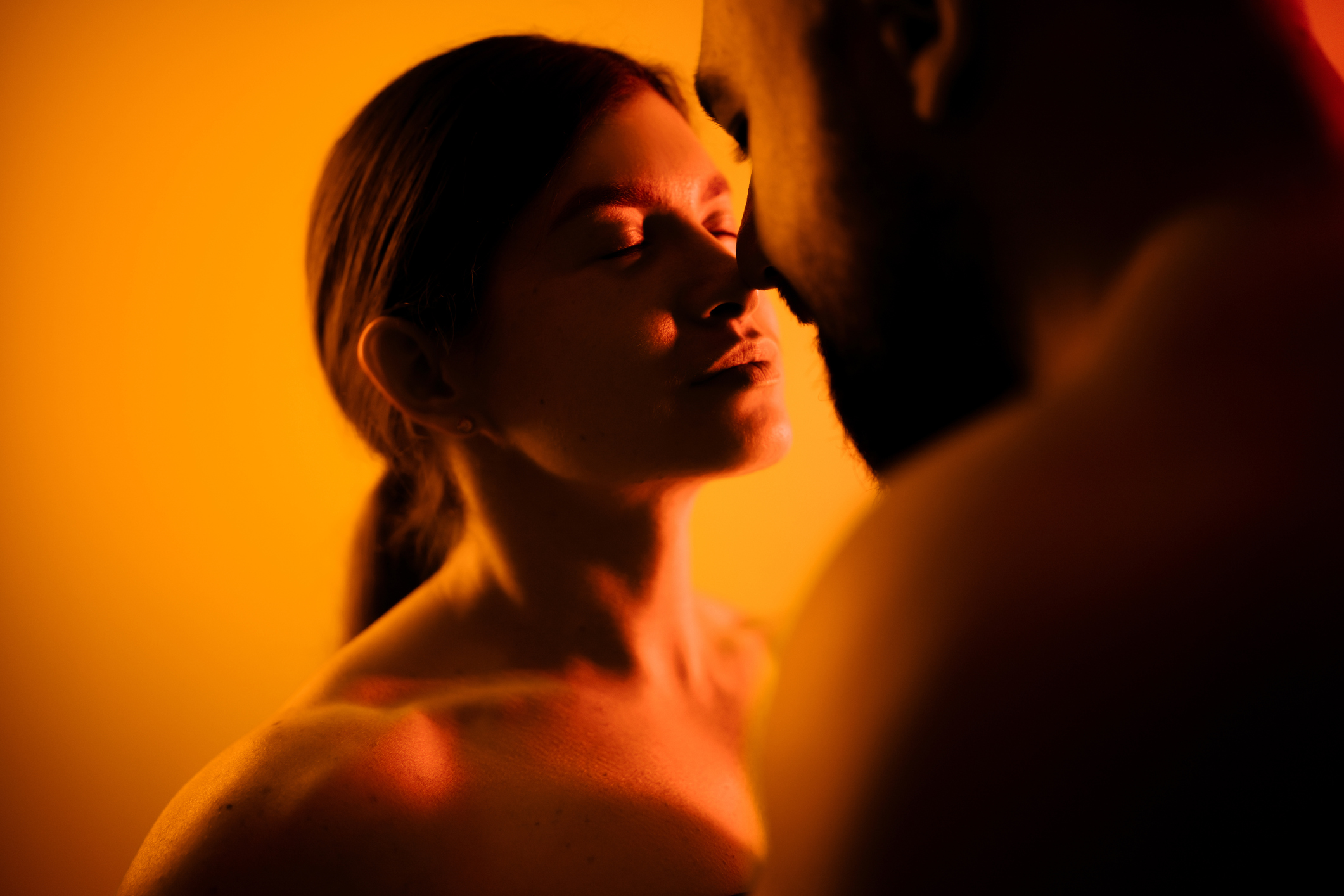 Close-up de um casal com a mulher de olhos fechados e o homem de costas, ambos iluminados por uma luz laranja e vermelha, criando um clima íntimo