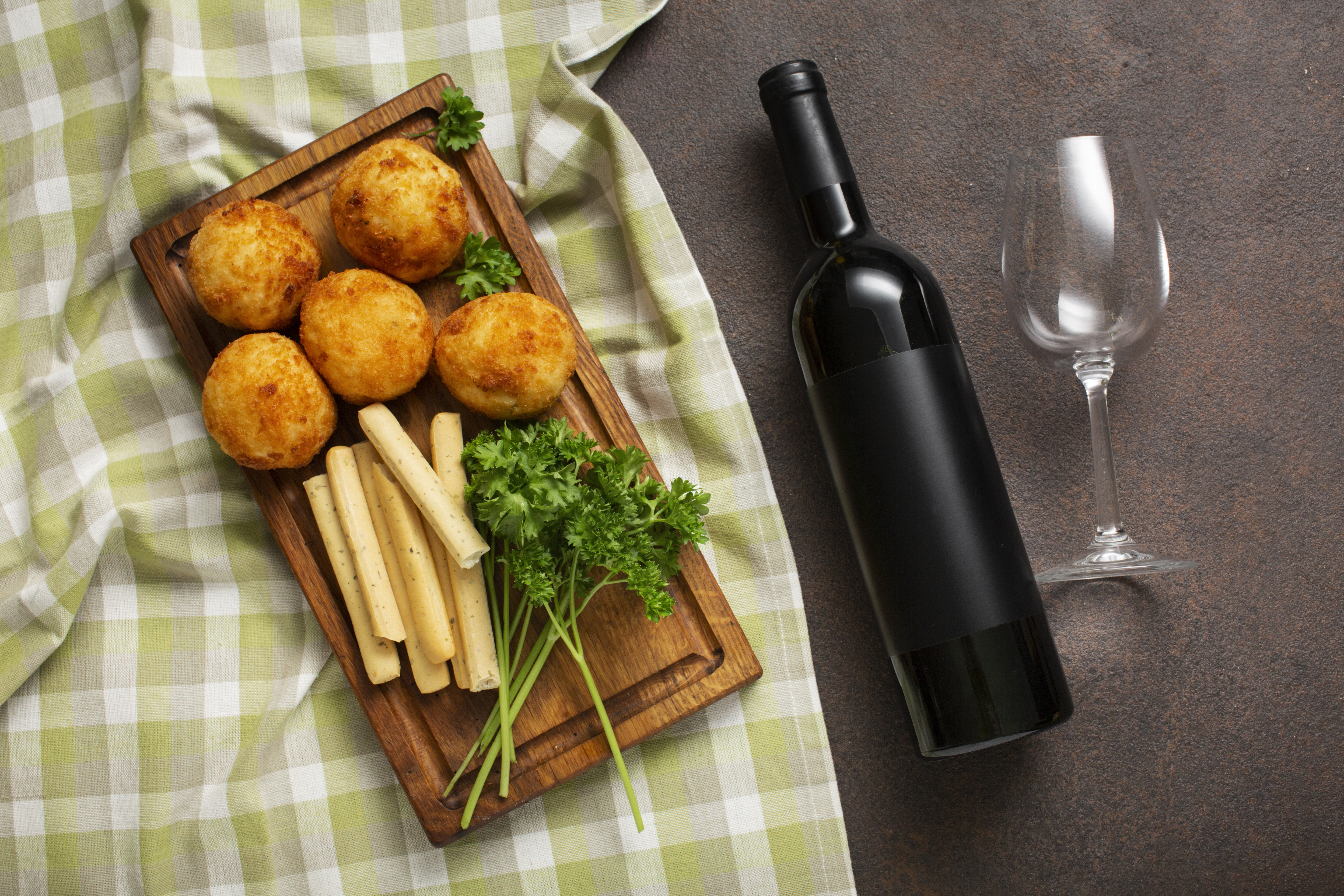 Uma garrafa de vinho tinto com rótulo preto, uma taça vazia e uma tábua de madeira com cinco bolinhos fritos dourados, palitos de pão e salsa fresca, sobre um pano de prato xadrez verde e branco, em uma superfície marrom escura