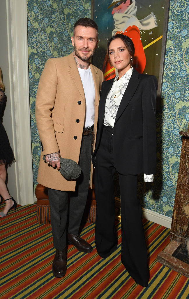David e Victoria Beckham posam sorridentes em um evento. Ele veste casaco bege, camiseta branca e cal&ccedil;a escura, segurando um bon&eacute;. Ela usa um blazer preto, cal&ccedil;a preta e blusa branca estampada. Ao fundo, papel de parede floral e um quadro com figura de esquiador. O ch&atilde;o tem um tapete listrado em tons de vermelho, amarelo e azul
