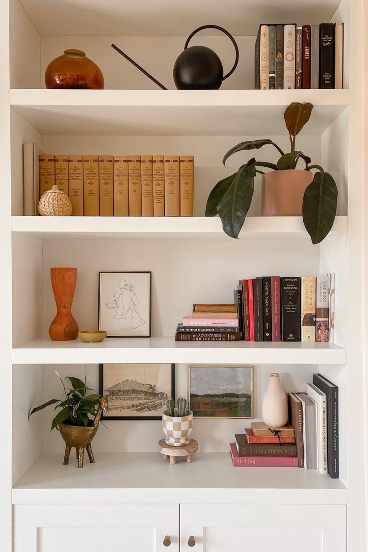 Estante branca com livros e objetos decorativos: vaso &acirc;mbar, regador preto, planta em vaso rosa, cestinha de palha, vaso de cer&acirc;mica laranja, tigela amarela, quadro com desenho abstrato, cacto em vaso xadrez, planta em vaso dourado de girafa, e pilhas de livros com capas variadas