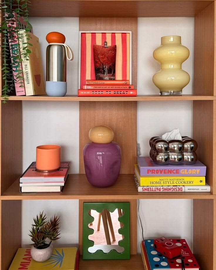 Estante de madeira clara com tr&ecirc;s prateleiras, exibindo objetos decorativos e livros coloridos. Na prateleira superior, uma garrafa t&eacute;rmica prateada com base azul e tampa laranja, um quadro com listras vermelhas e laranja e uma imagem de bebida, e um vaso amarelo claro com formato org&acirc;nico. Na prateleira do meio, um copo laranja sobre livros, um vaso roxo com tampa bege, e uma caixa de len&ccedil;os cromada sobre livros. Na prateleira inferior, um pequeno vaso com planta suculenta, um espelho verde com bordas onduladas, e uma c&acirc;mera vermelha sobre livros coloridos