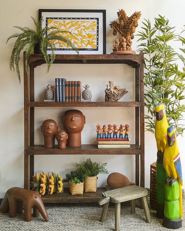 Estante de madeira r&uacute;stica com objetos de arte popular brasileira, incluindo xilogravura de p&aacute;ssaros, esculturas de barro, livros e plantas, ao lado de uma planta alta e arara de madeira amarela e azul