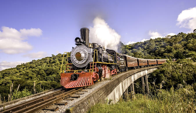 Locomotiva a vapor preta e vermelha com fuma&ccedil;a branca saindo da chamin&eacute;, puxando vag&otilde;es marrons sobre uma ponte de concreto, cercada por colinas verdes e c&eacute;u azul com nuvens esparsas