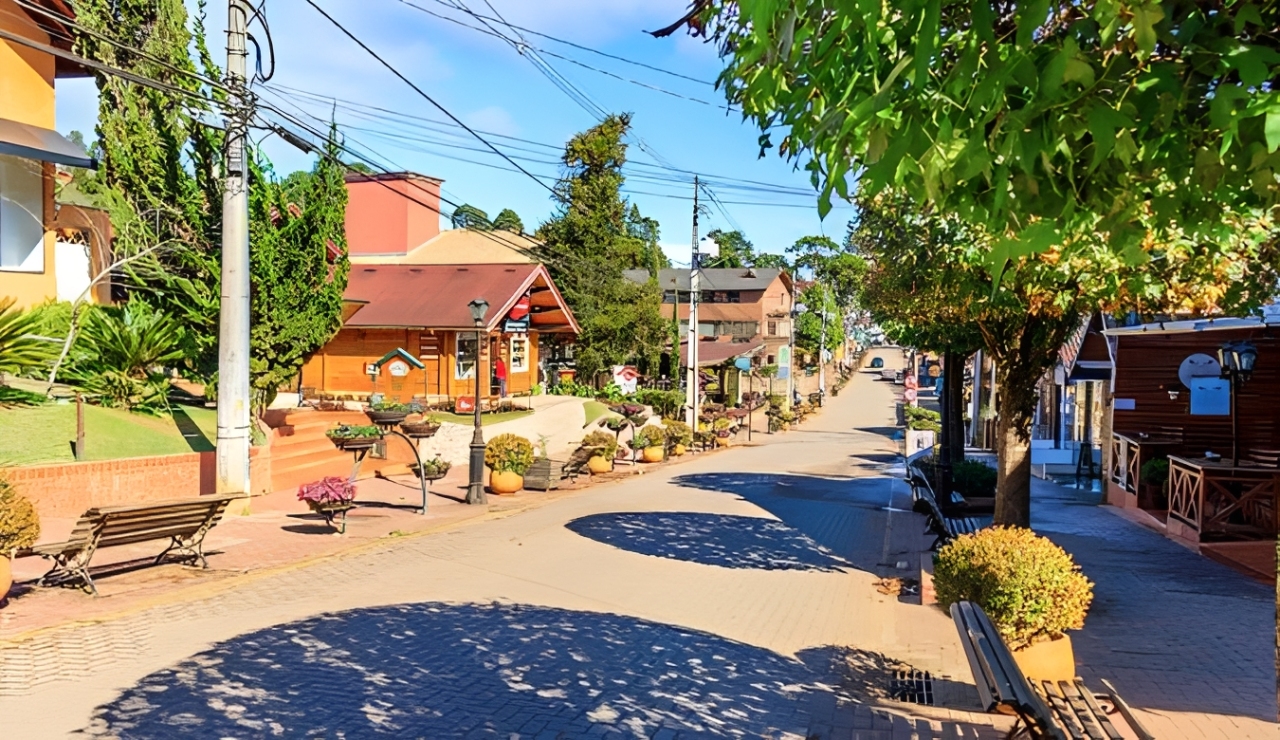 Rua pavimentada com paralelep&iacute;pedos, ladeada por casas de madeira e telhados marrons, bancos de madeira e vasos de flores coloridas. &Aacute;rvores frondosas projetam sombras no ch&atilde;o, sob um c&eacute;u azul claro com poucas nuvens. Postes com fia&ccedil;&atilde;o el&eacute;trica vis&iacute;veis.
