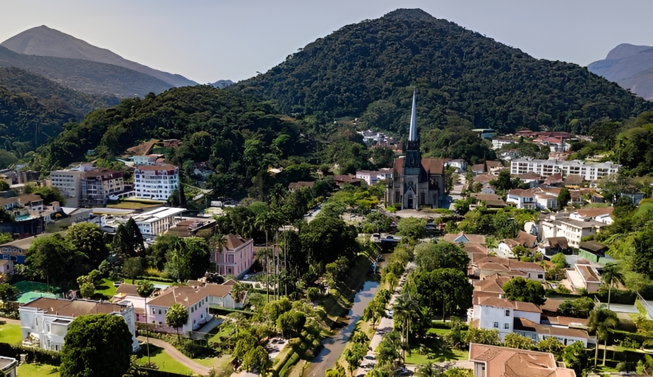 Vista a&eacute;rea de uma cidade com uma grande igreja g&oacute;tica de torre pontiaguda no centro, cercada por casas e pr&eacute;dios baixos, com muitas &aacute;rvores e montanhas verdes ao fundo, sob um c&eacute;u azul claro