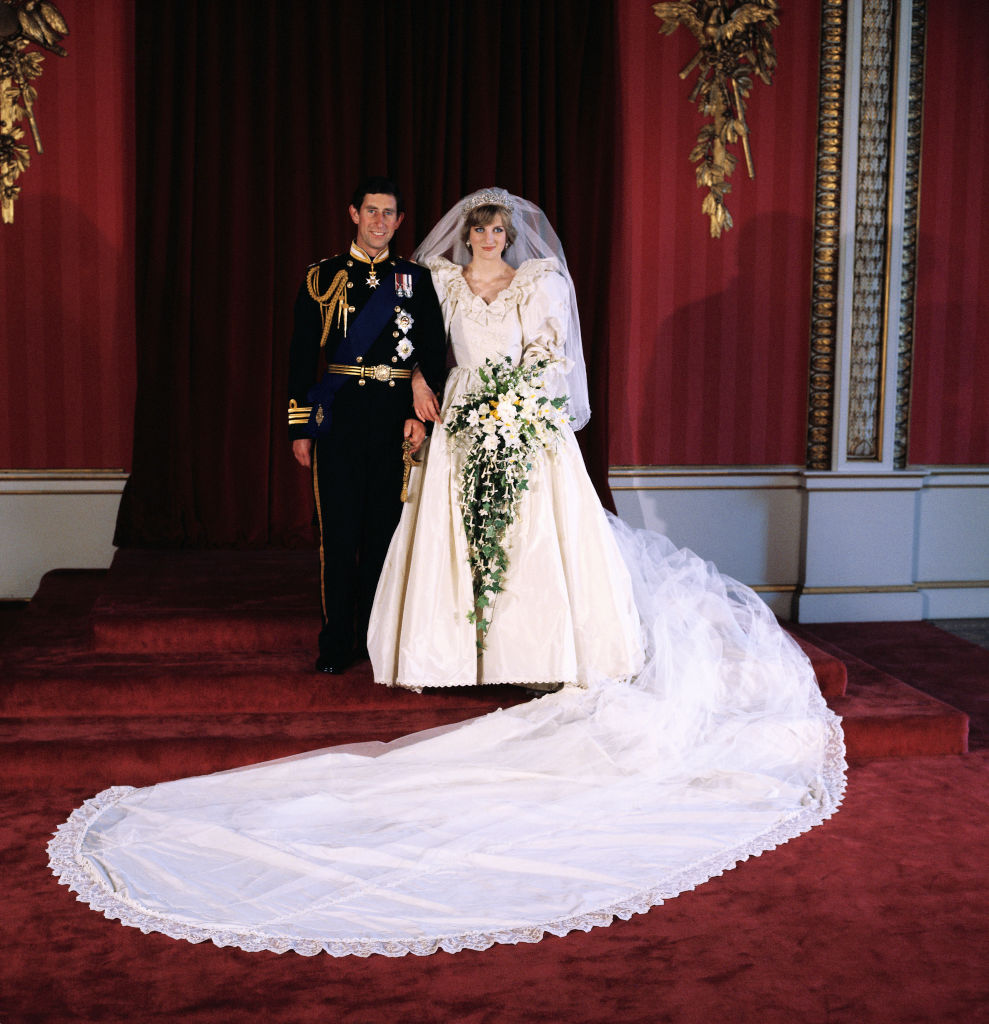 Pr&iacute;ncipe Charles e Princesa Diana posam para foto de casamento. Charles veste uniforme militar azul-marinho com medalhas, e Diana usa um vestido de noiva branco volumoso com mangas bufantes, v&eacute;u e uma cauda longa de renda que se estende pelo tapete vermelho. Ela segura um buqu&ecirc; cascata de flores brancas e folhagens verdes. Ambos sorriem levemente, em p&eacute; sobre degraus forrados com carpete vermelho, em um ambiente com cortinas e detalhes dourados