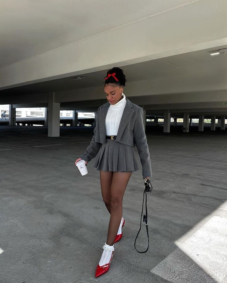 Mulher jovem com cabelo preso e la&ccedil;o vermelho, vestindo su&eacute;ter branco, blazer cinza cropped, saia plissada cinza, meia-cal&ccedil;a escura, meias brancas de renda e sapatos vermelhos de salto, caminhando em estacionamento vazio com um copo na m&atilde;o e bolsa preta pendurada no bra&ccedil;o