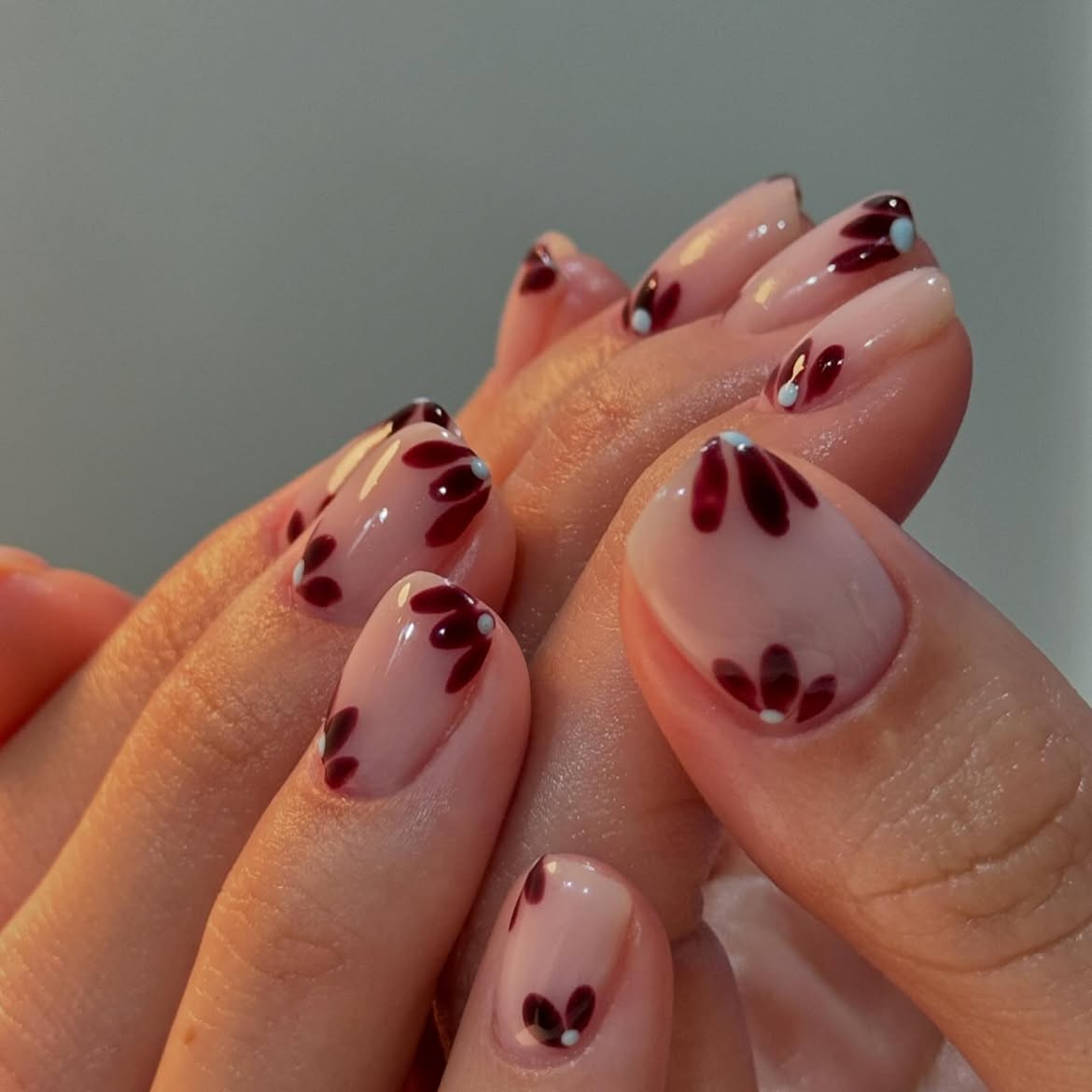 M&atilde;os com unhas curtas e arredondadas, esmalte nude com desenhos de flores em vinho e miolo branco, e pontos azuis claros em algumas unhas