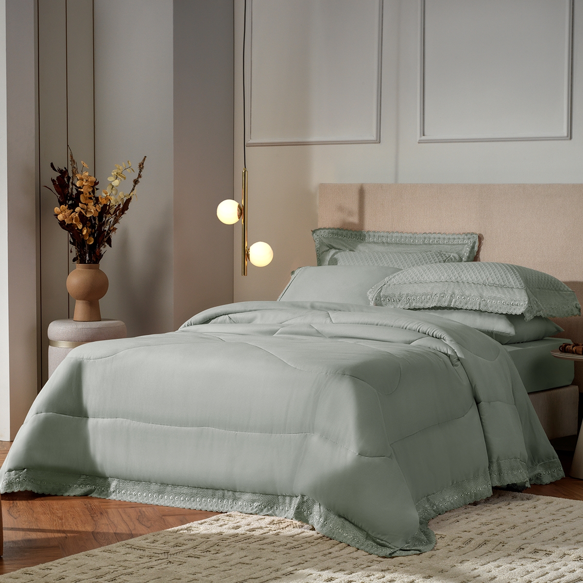 Cama de casal com edredom e travesseiros verde-&aacute;gua, detalhes de renda na barra. &Agrave; esquerda, vaso de cer&acirc;mica com flores secas sobre uma mesa de cabeceira redonda. Pendente dourado com duas esferas de luz ilumina o ambiente. O ch&atilde;o &eacute; de madeira com um tapete bege felpudo. A parede tem molduras decorativas