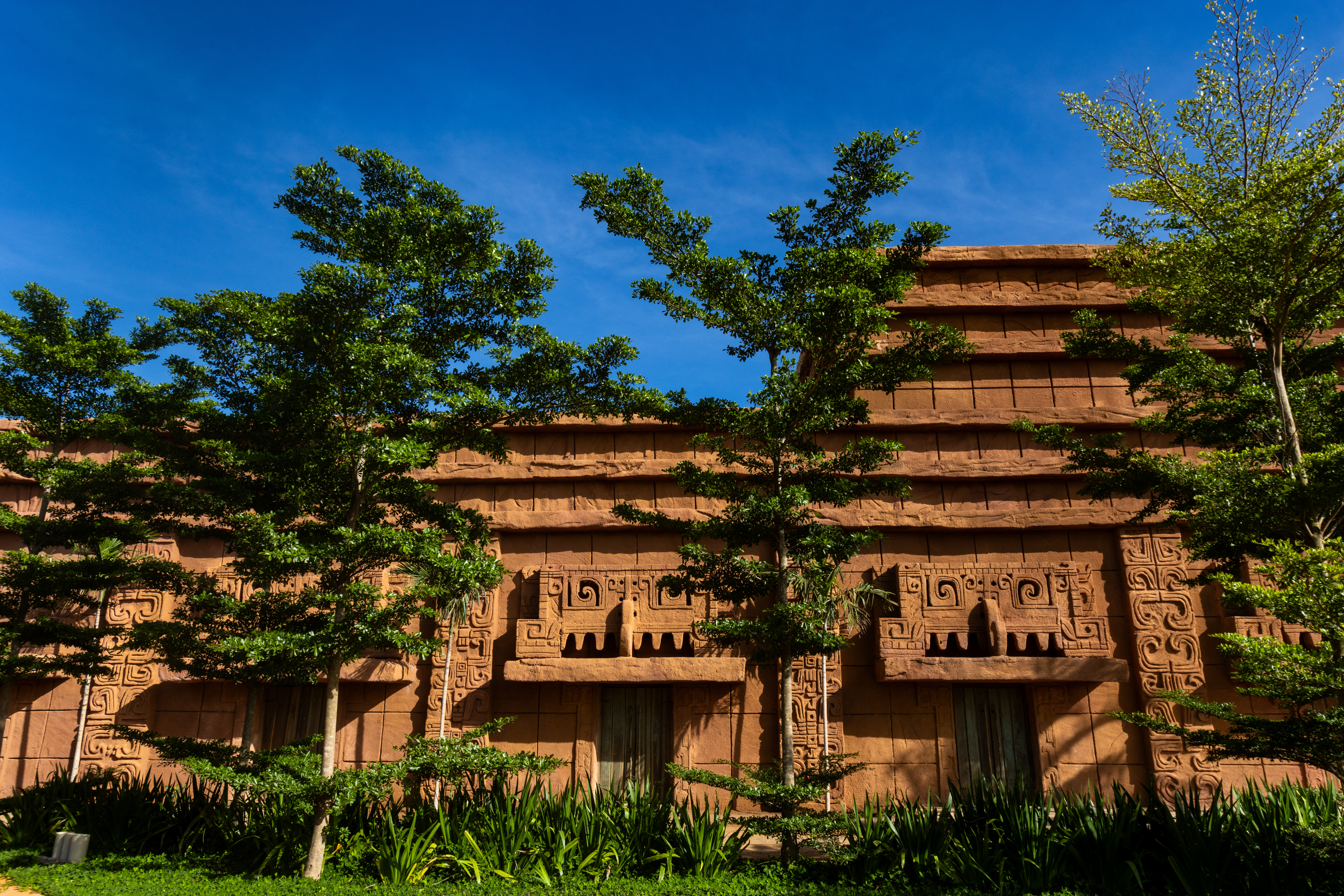 Fachada de edifício em estilo maia, com paredes de cor terracota e relevos geométricos, sob céu azul. Árvores de folhas verdes e arbustos cobrem parte da estrutura, com portas escuras visíveis na base