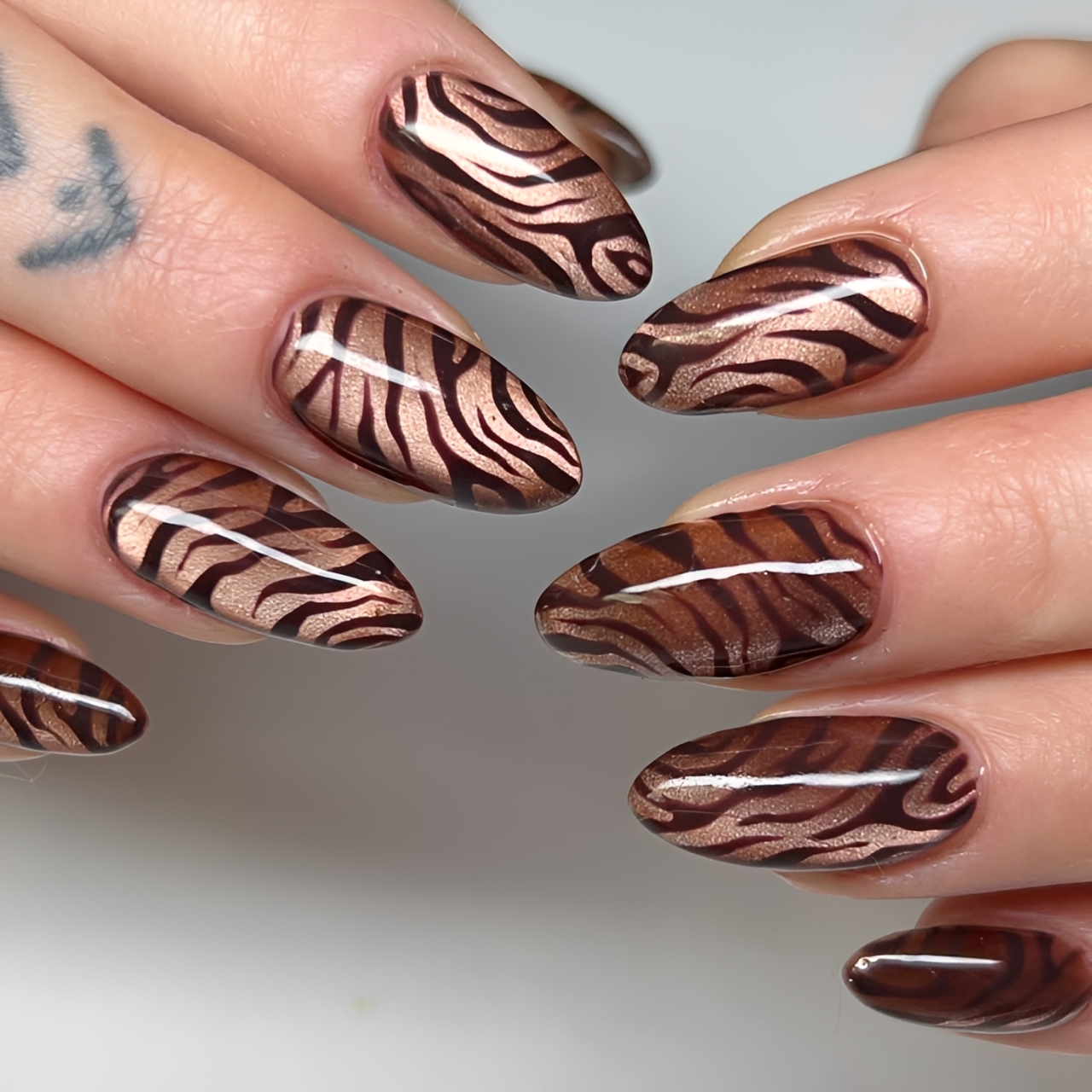M&atilde;os com unhas ovais longas, esmaltadas em marrom escuro com listras onduladas em tom bronze met&aacute;lico, imitando estampa de tigre