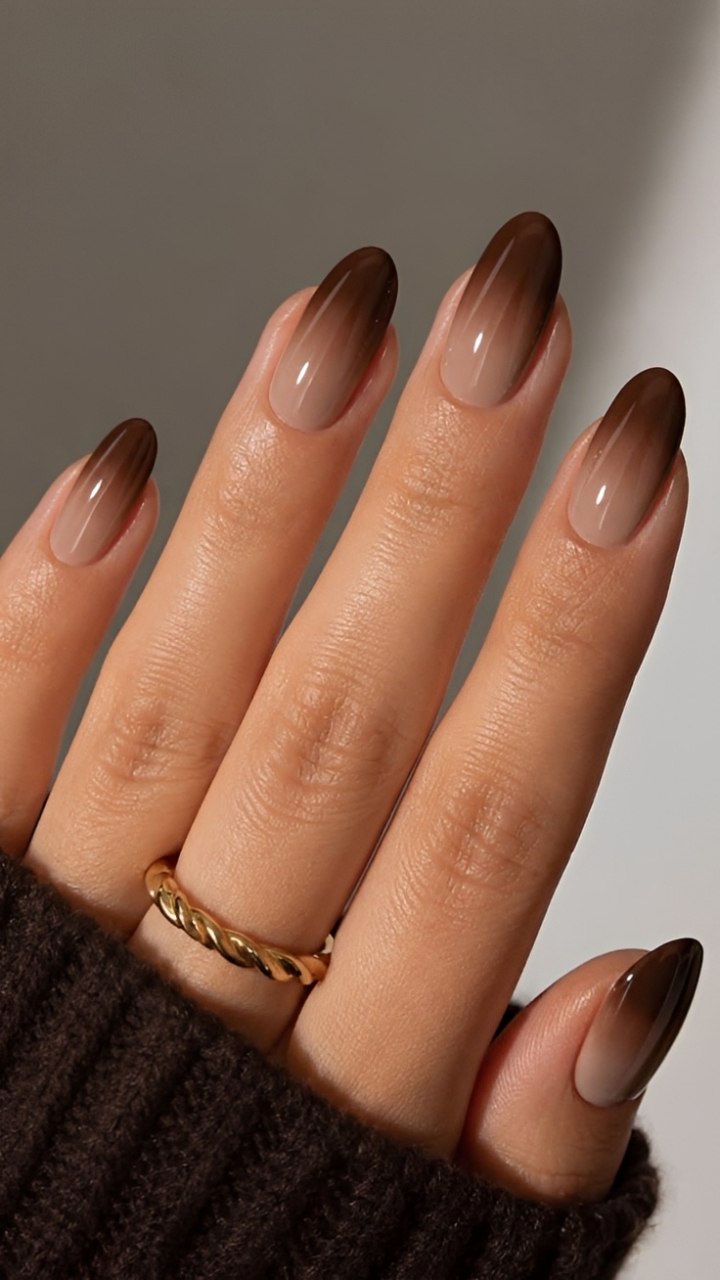 M&atilde;o feminina com unhas ovais longas em ombr&eacute;, degrad&ecirc; do nude claro na base para o marrom escuro nas pontas. Um anel dourado torcido adorna o dedo anelar. A m&atilde;o veste uma manga de su&eacute;ter marrom escuro