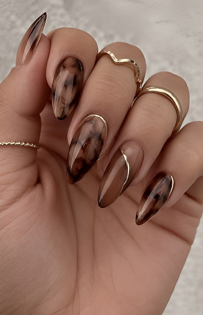 M&atilde;o feminina com unhas amendoadas longas, esmalte marrom com efeito tartaruga e detalhes dourados, usando an&eacute;is finos dourados