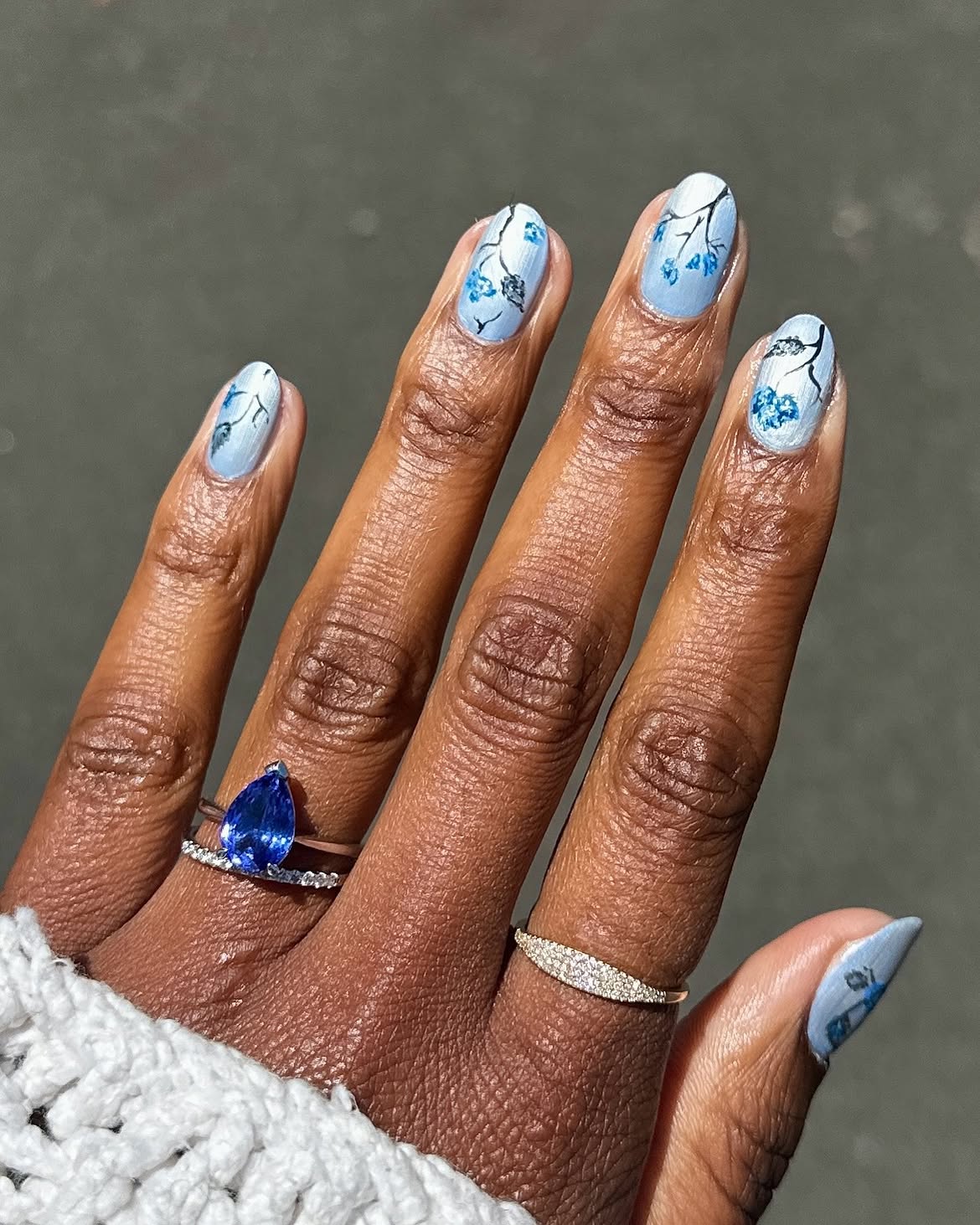 M&atilde;o de pele escura com unhas ovais pintadas de azul claro, decoradas com galhos pretos e flores azuis. Dois an&eacute;is de prata adornam os dedos: um com safira azul em formato de gota e outro com diamantes
