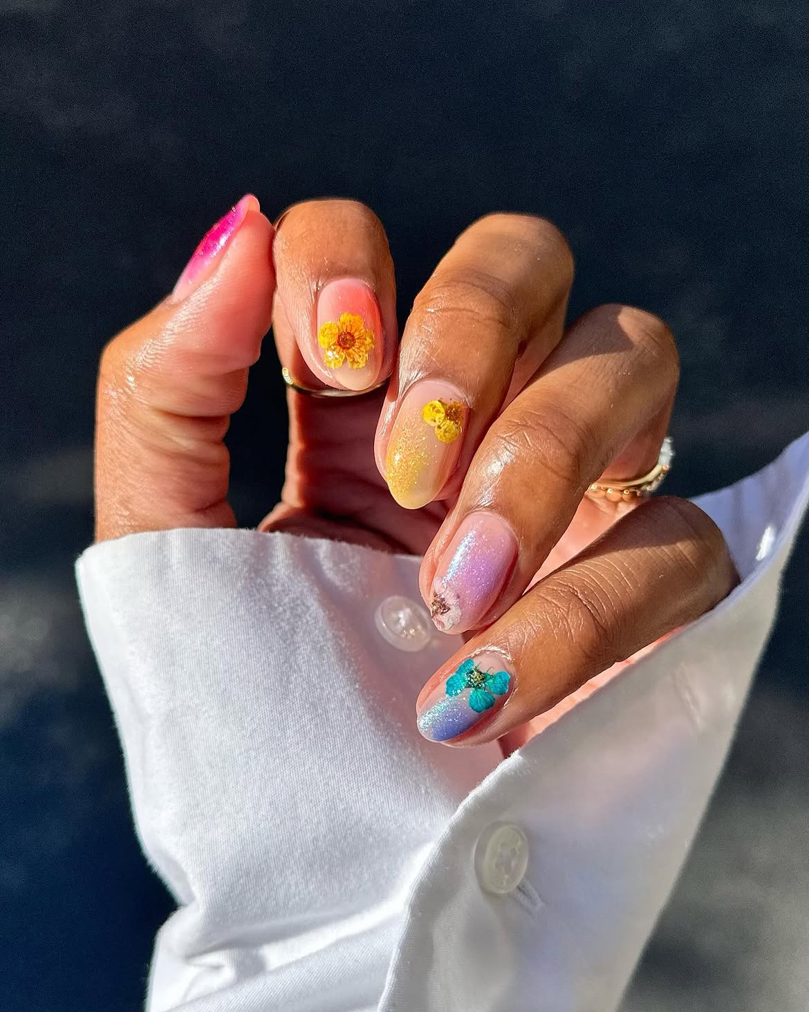 M&atilde;o com unhas decoradas com flores secas. O polegar tem esmalte rosa vibrante, o indicador e o m&eacute;dio t&ecirc;m esmalte degrad&ecirc; de p&ecirc;ssego a amarelo com flores amarelas. O anelar tem esmalte lil&aacute;s com uma flor branca e o mindinho tem esmalte azul e lil&aacute;s com uma flor azul. A m&atilde;o veste uma camisa branca de manga longa