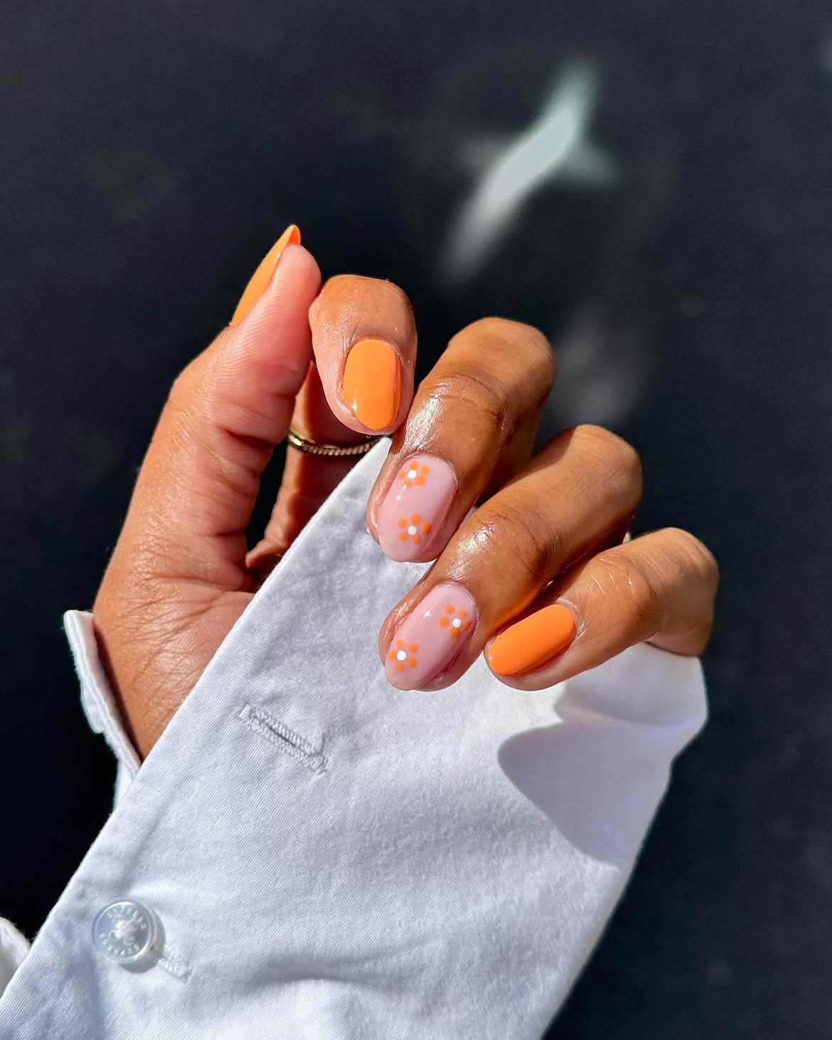 M&atilde;o de pele morena com unhas curtas e arredondadas. O polegar e o dedo m&iacute;nimo t&ecirc;m esmalte laranja vibrante, enquanto os dedos indicador, m&eacute;dio e anelar exibem esmalte rosa claro com pequenas flores laranjas e pontos brancos no centro. A m&atilde;o segura um tecido branco de camisa com um bot&atilde;o vis&iacute;vel, sobre um fundo escuro e desfocado