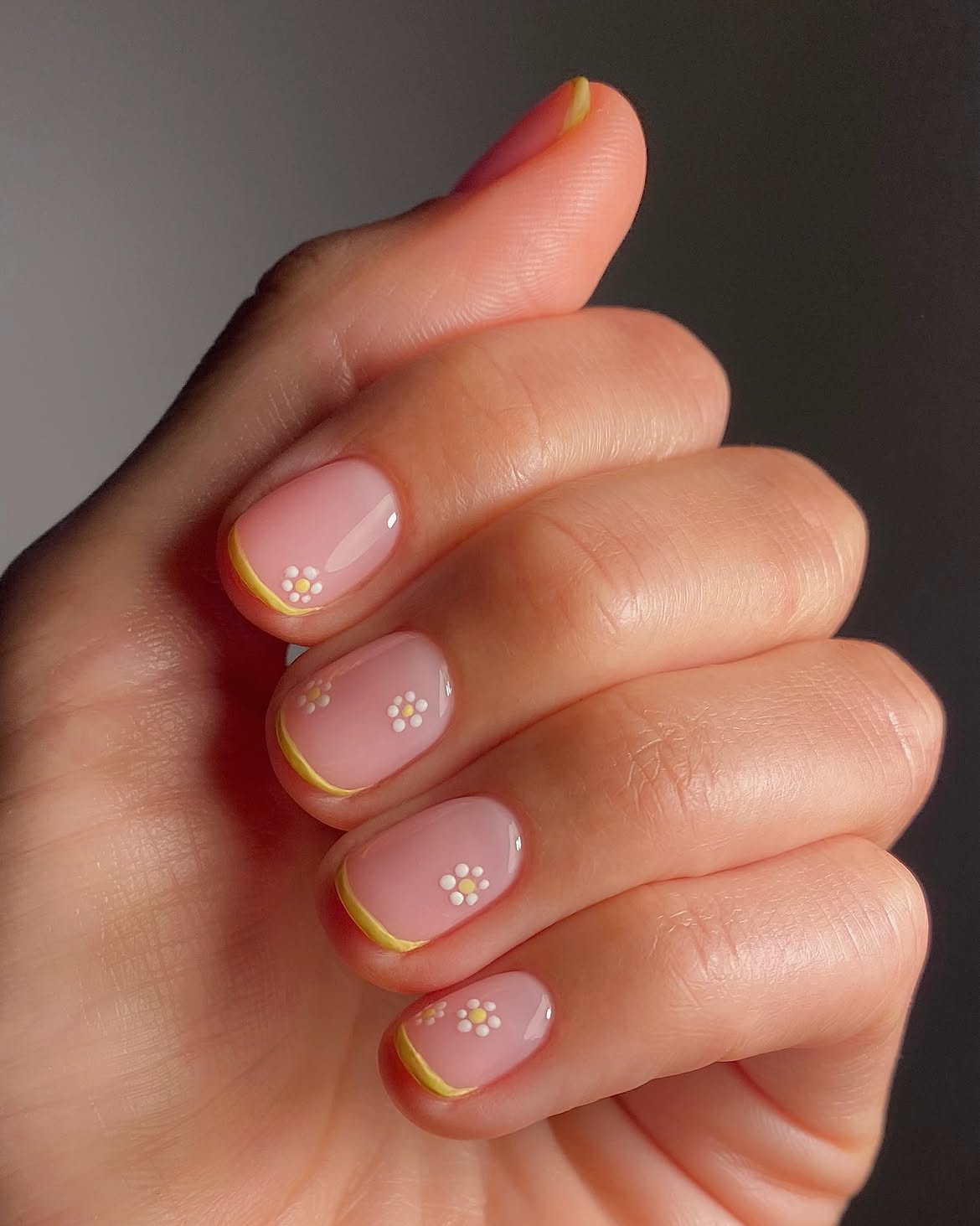 M&atilde;o feminina com unhas curtas pintadas de rosa claro, com pontas amarelas e pequenas flores brancas com miolo amarelo em cada unha