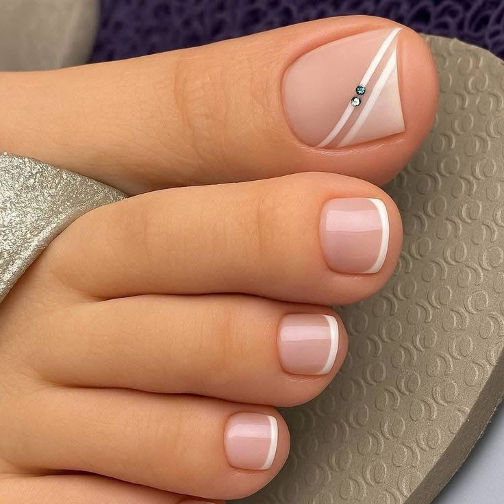 P&eacute;s femininos com unhas dos dedos pintadas em estilo francesinha, com base nude e pontas brancas. O ded&atilde;o tem um design diagonal com duas linhas brancas e dois pequenos strass azuis, usando um chinelo cinza claro com textura