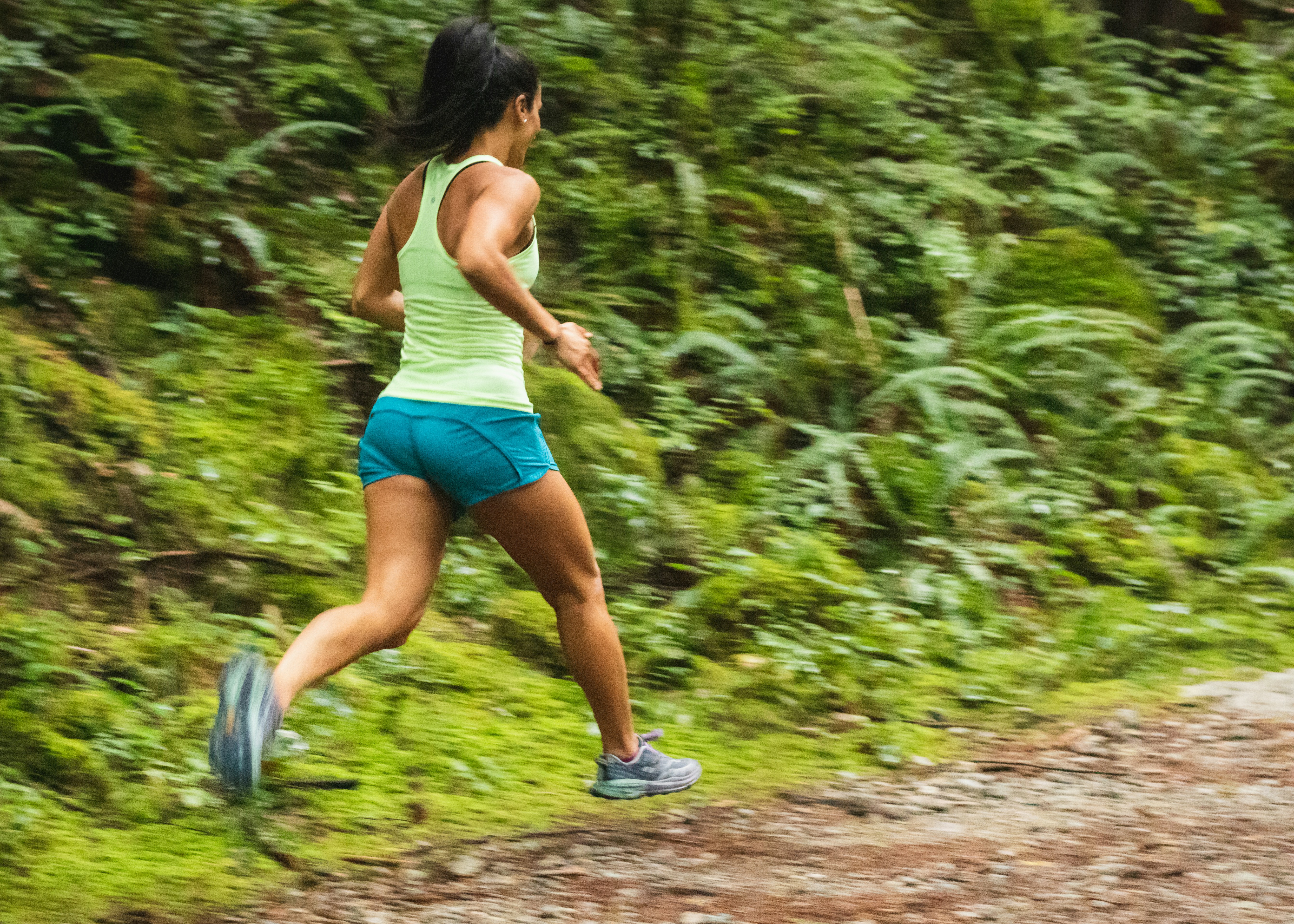 Mulher de costas, com rabo de cavalo, camiseta verde-lim&atilde;o e shorts azul-turquesa, correndo em trilha de terra com vegeta&ccedil;&atilde;o verde exuberante ao fundo