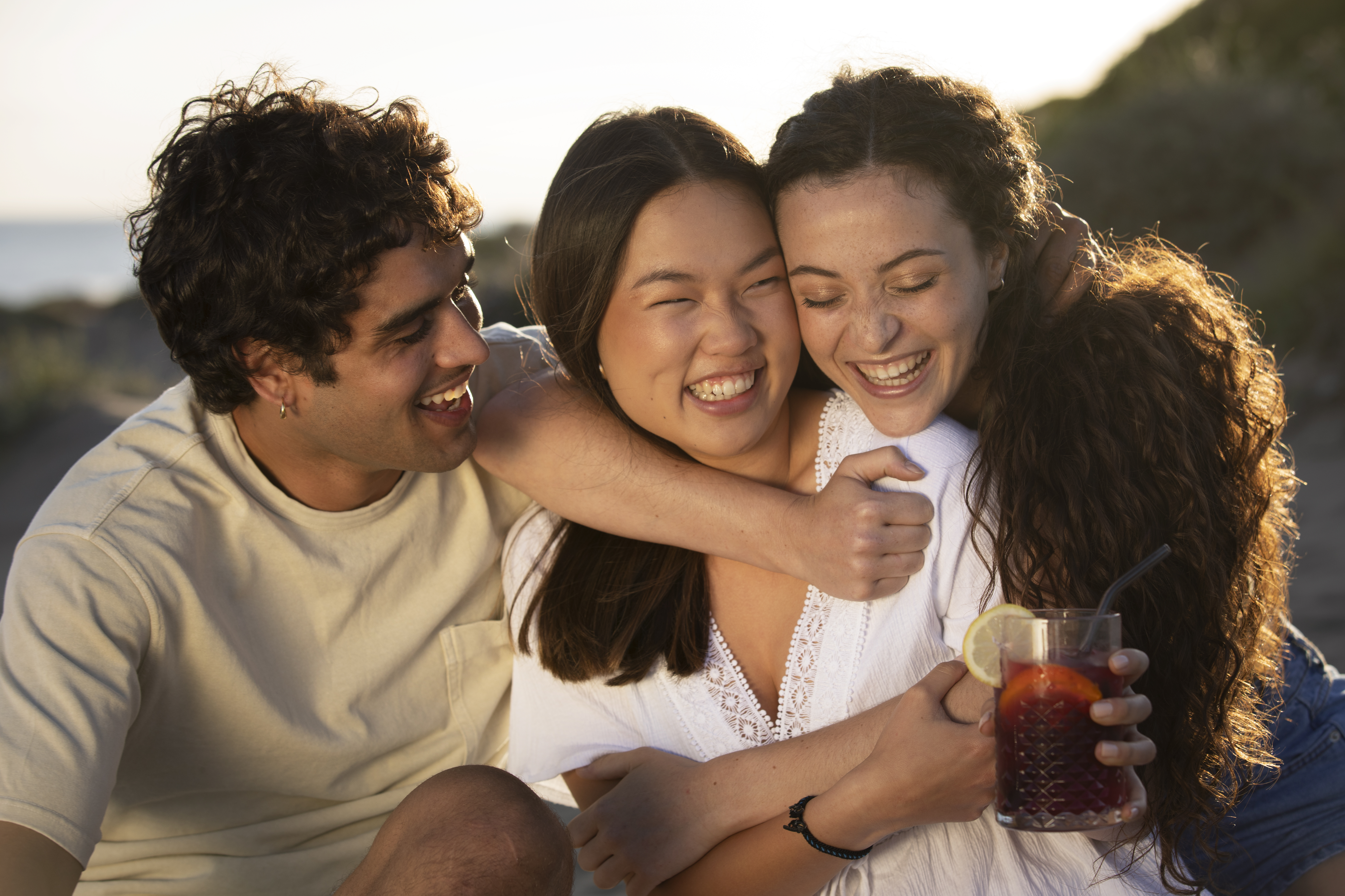 Tr&ecirc;s amigos sorrindo e abra&ccedil;ando ao ar livre. Um homem &agrave; esquerda, de camiseta bege, abra&ccedil;a duas mulheres. A mulher do centro, de blusa branca, tem os olhos fechados e sorri. A mulher da direita, de blusa branca e cabelo cacheado, tamb&eacute;m sorri com os olhos fechados e segura um copo de bebida vermelha com lim&atilde;o